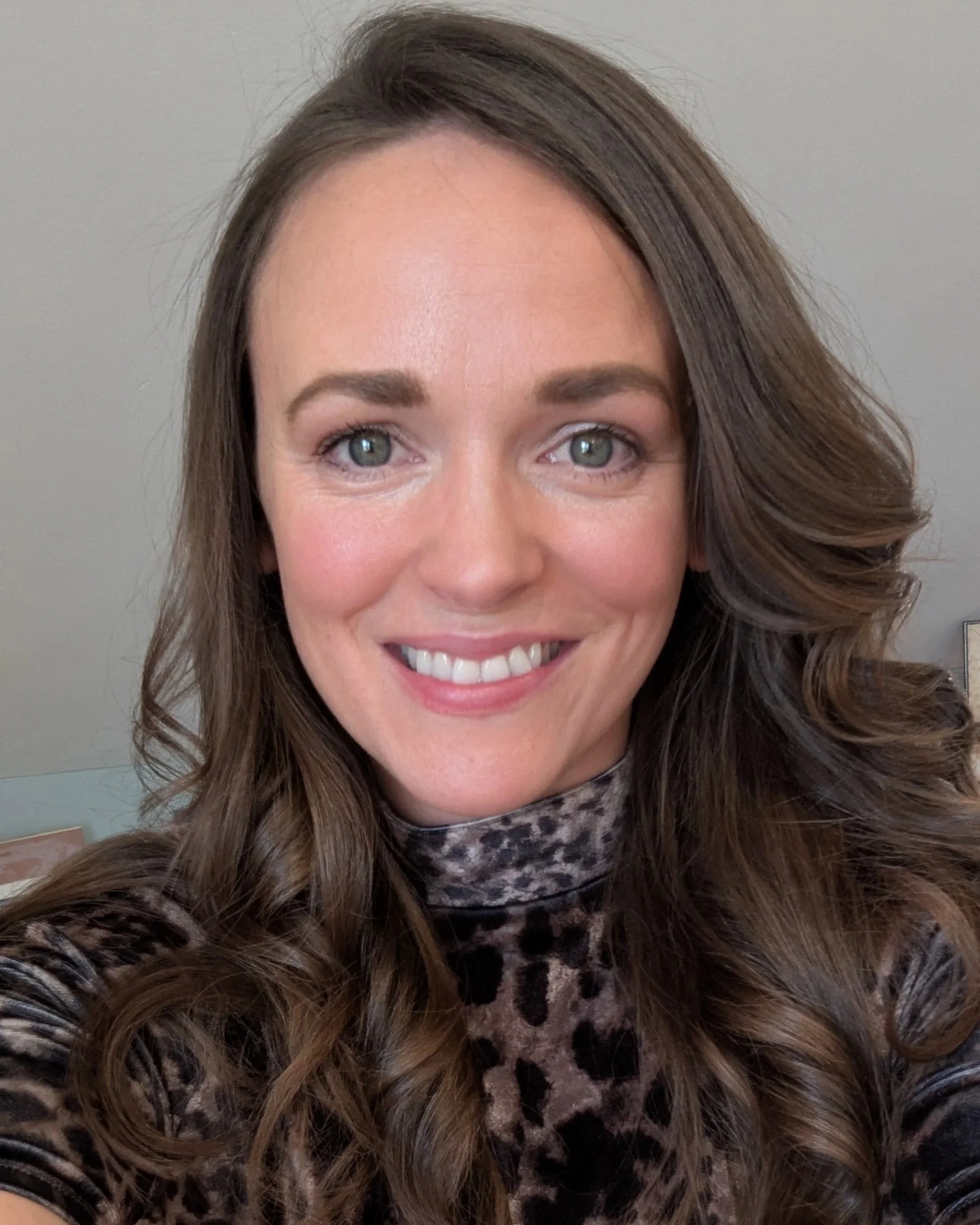 A woman with long brown curly hair, wearing a leopard print top, smiling at the camera.