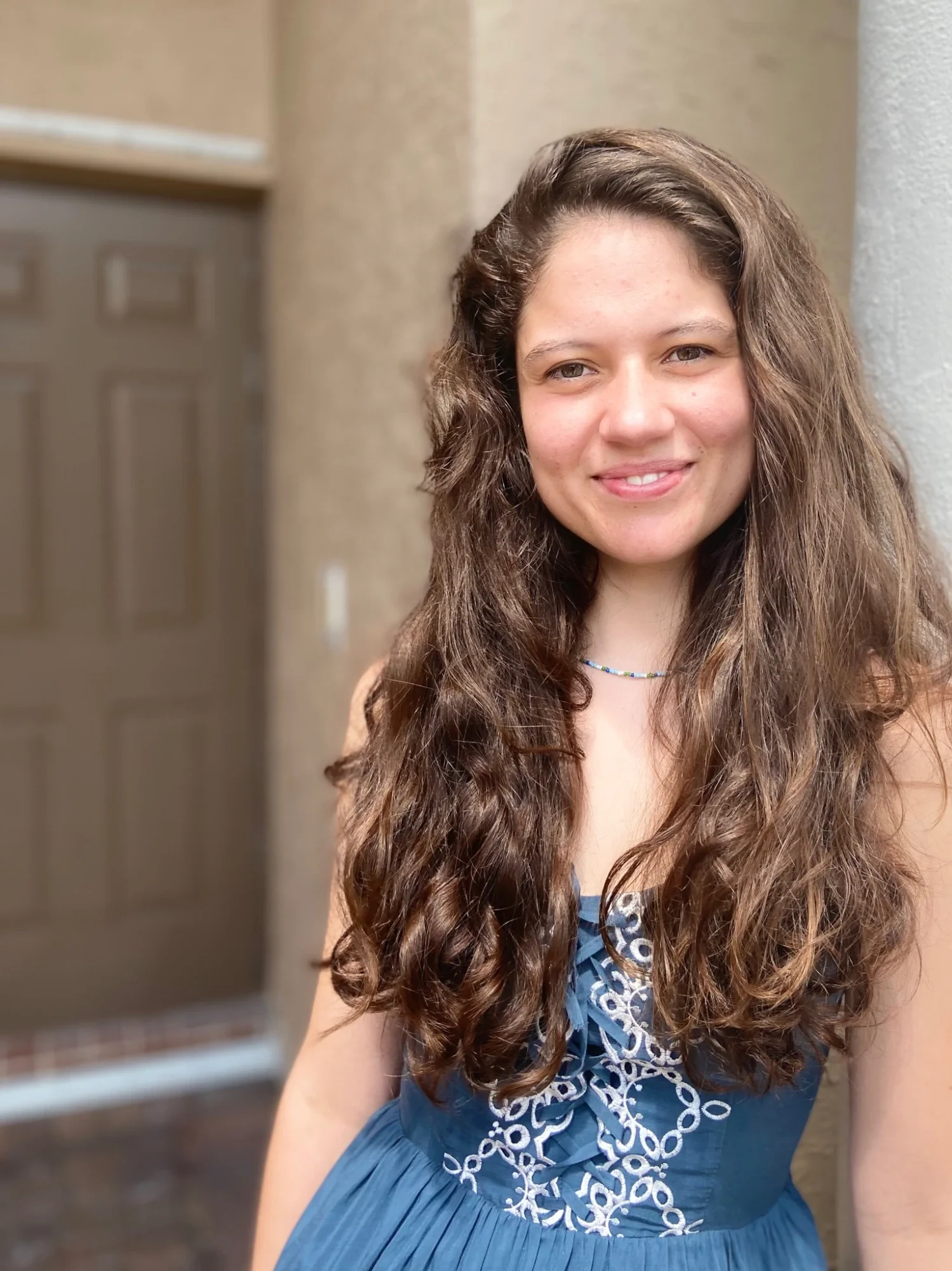 A young woman with long, wavy brown hair, smiling, wearing a blue dress with embroidery and a beaded necklace, standing outdoors near a building.
