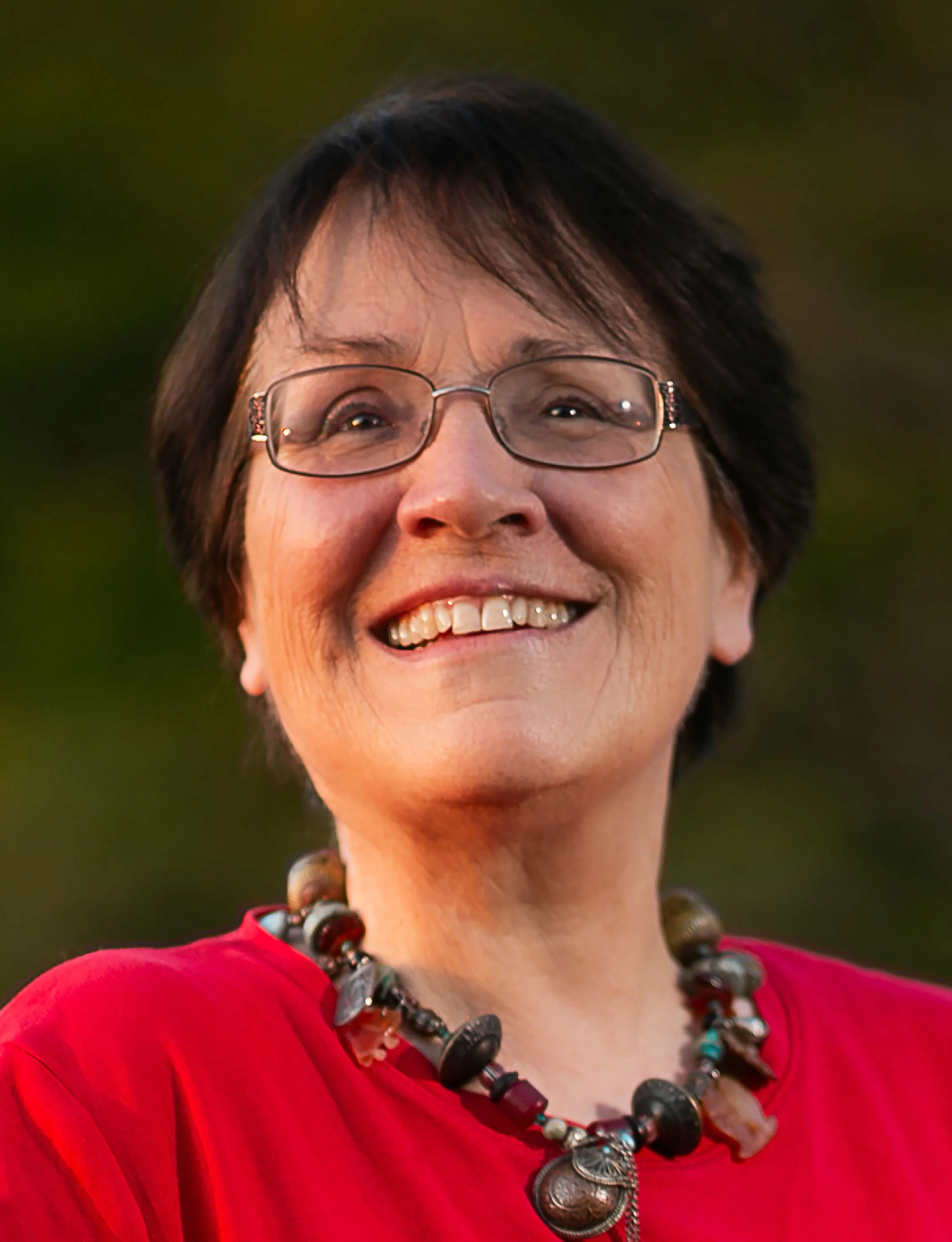 Portrait of a smiling woman with short dark hair, glasses, wearing a red top and a colorful beaded necklace.