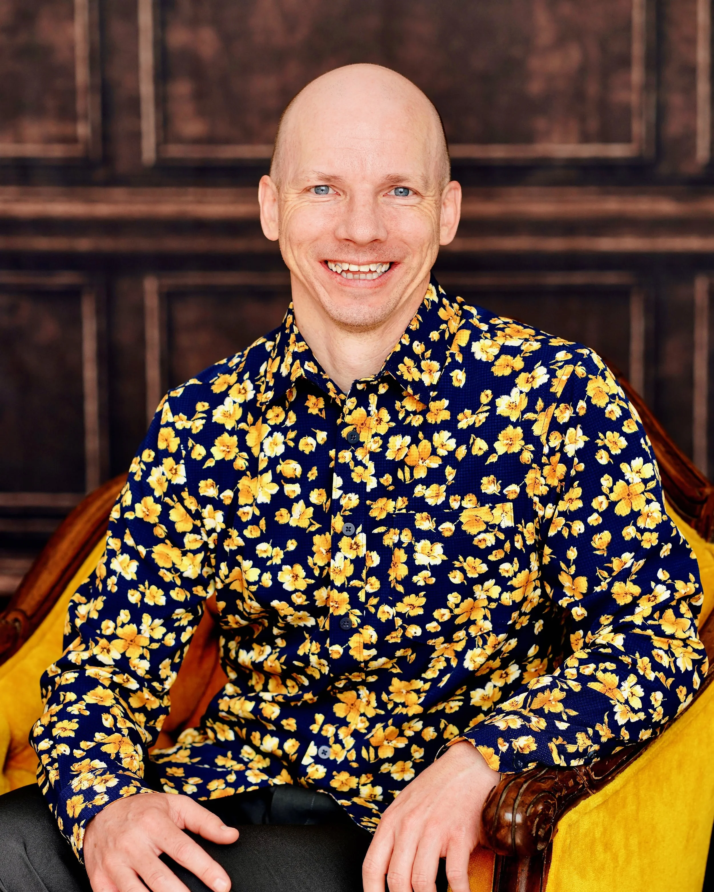 A smiling man sitting in a yellow armchair with a wooden frame, wearing a dark blue shirt with a yellow floral pattern, in front of a wooden paneled background.
