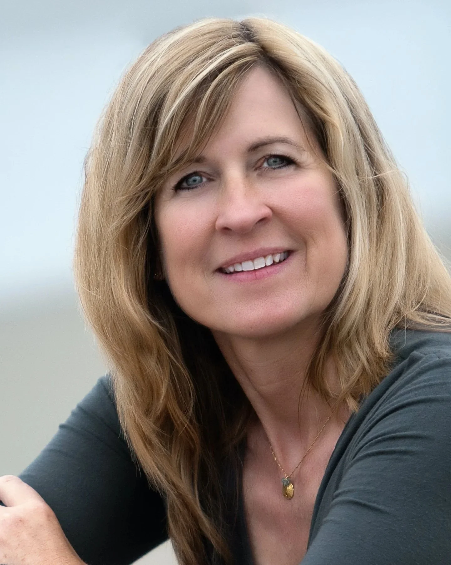 A woman with shoulder-length light brown hair and blue eyes smiling outdoors with a blurred background.
