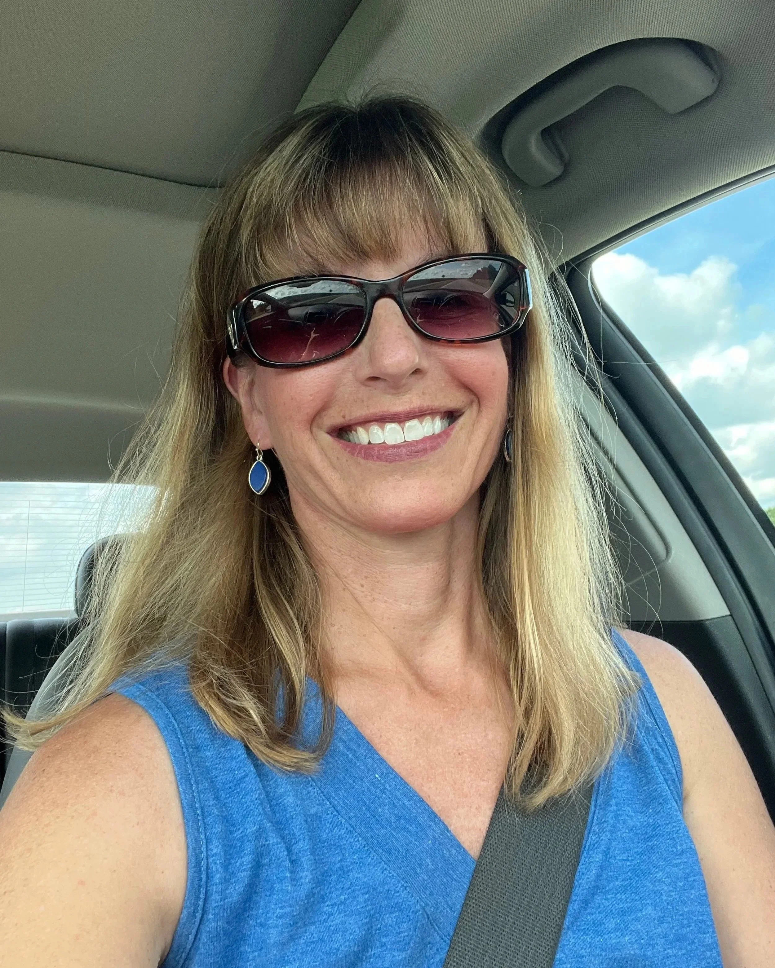 A smiling woman with blonde hair and sunglasses sitting in a car, wearing a blue sleeveless top, with a sunny sky visible outside the window.