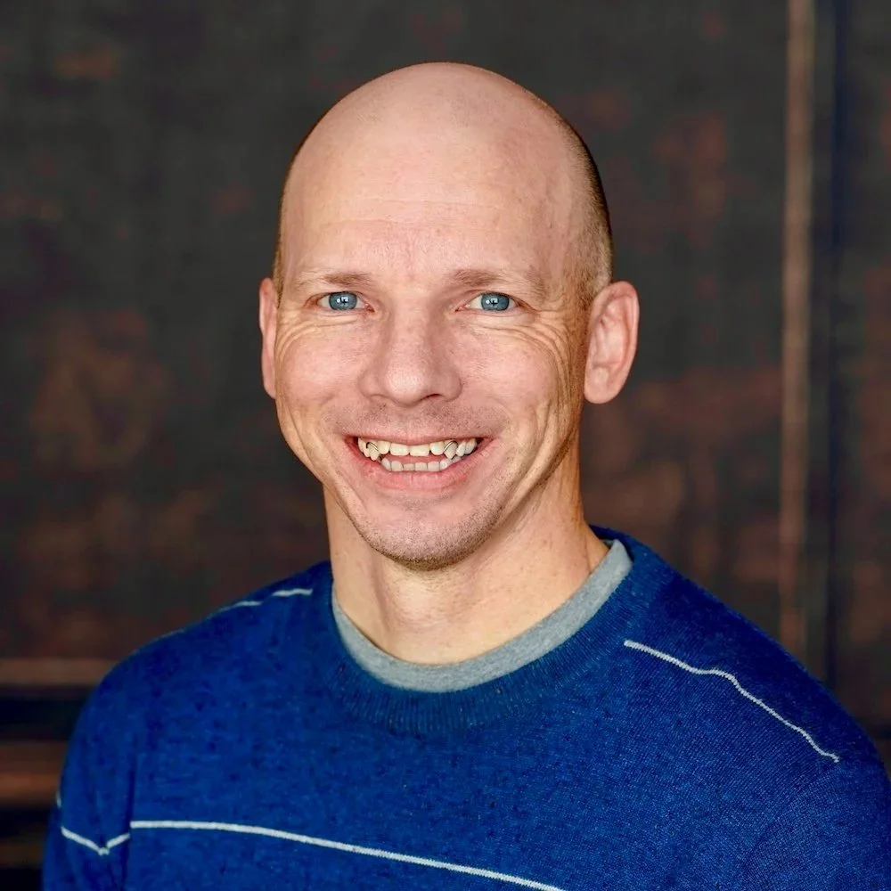 A smiling man with a shaved head wearing a black checkered shirt against a plain white background.