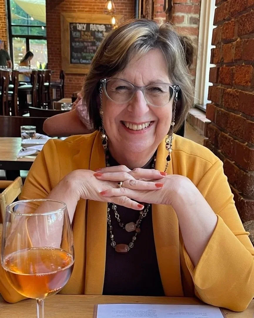 Smiling woman with glasses and jewelry sitting at a restaurant table, with a glass of rosé wine in front of her and a brick wall in the background.