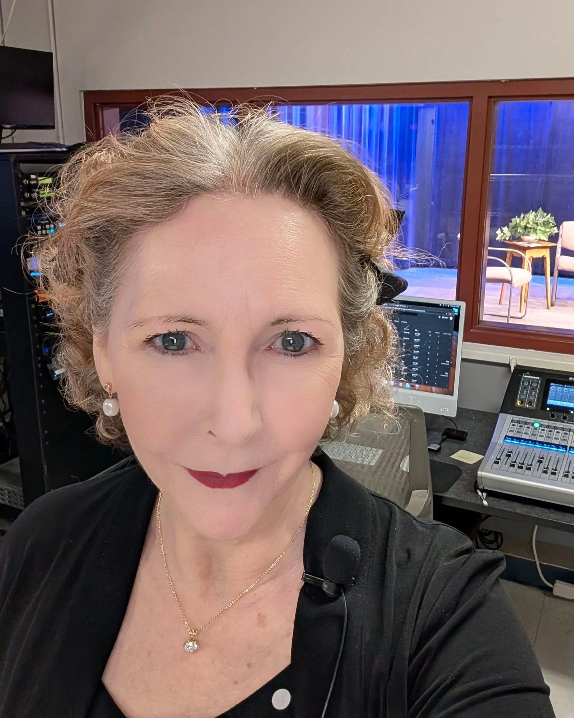 Close-up selfie of a middle-aged woman with curly blonde hair, wearing pearl earrings and a necklace, in a recording studio with audio equipment and a window showing a seating area with chairs and a table in the background.