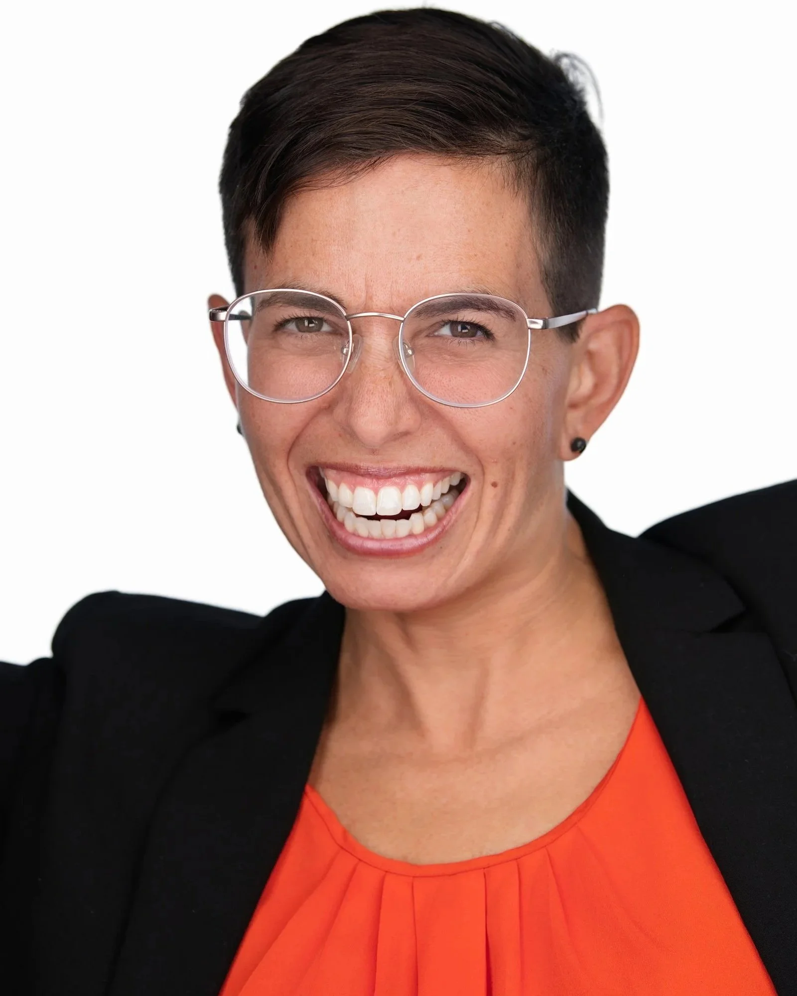 A woman with short dark hair wearing glasses, a black blazer, and an orange top, smiling broadly against a plain white background.
