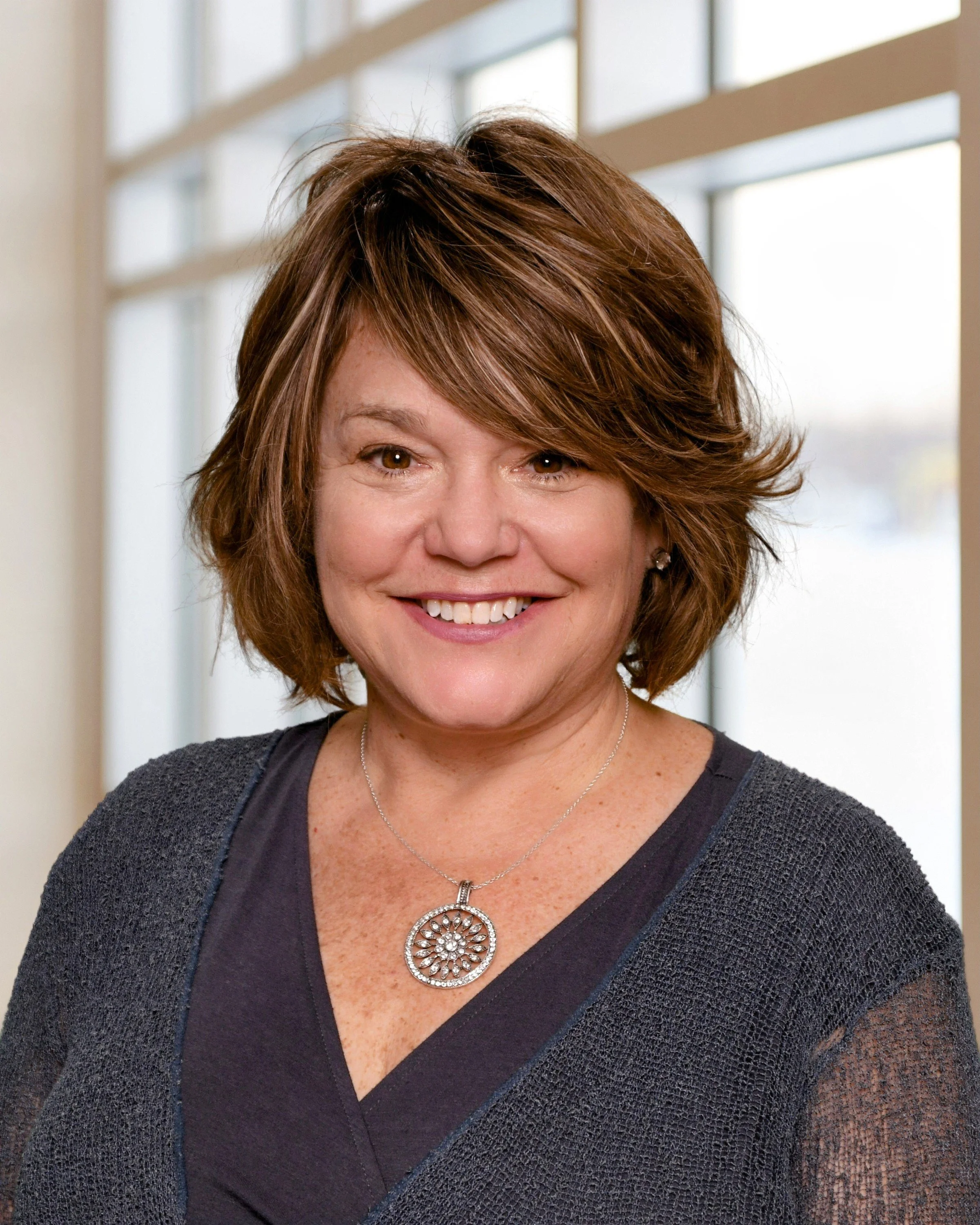 A smiling woman with brown hair, wearing a dark top with a sheer jacket and a silver necklace, standing in front of a large window.