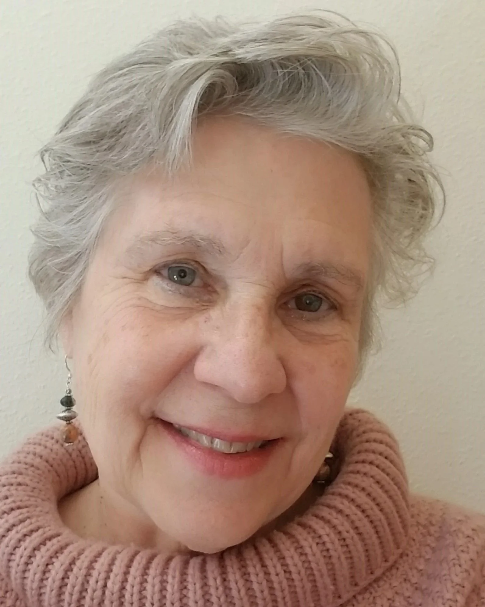 Close-up photo of a smiling elderly woman with short, gray hair, wearing a pink knitted turtleneck sweater and dangling earrings, against a light-colored background.