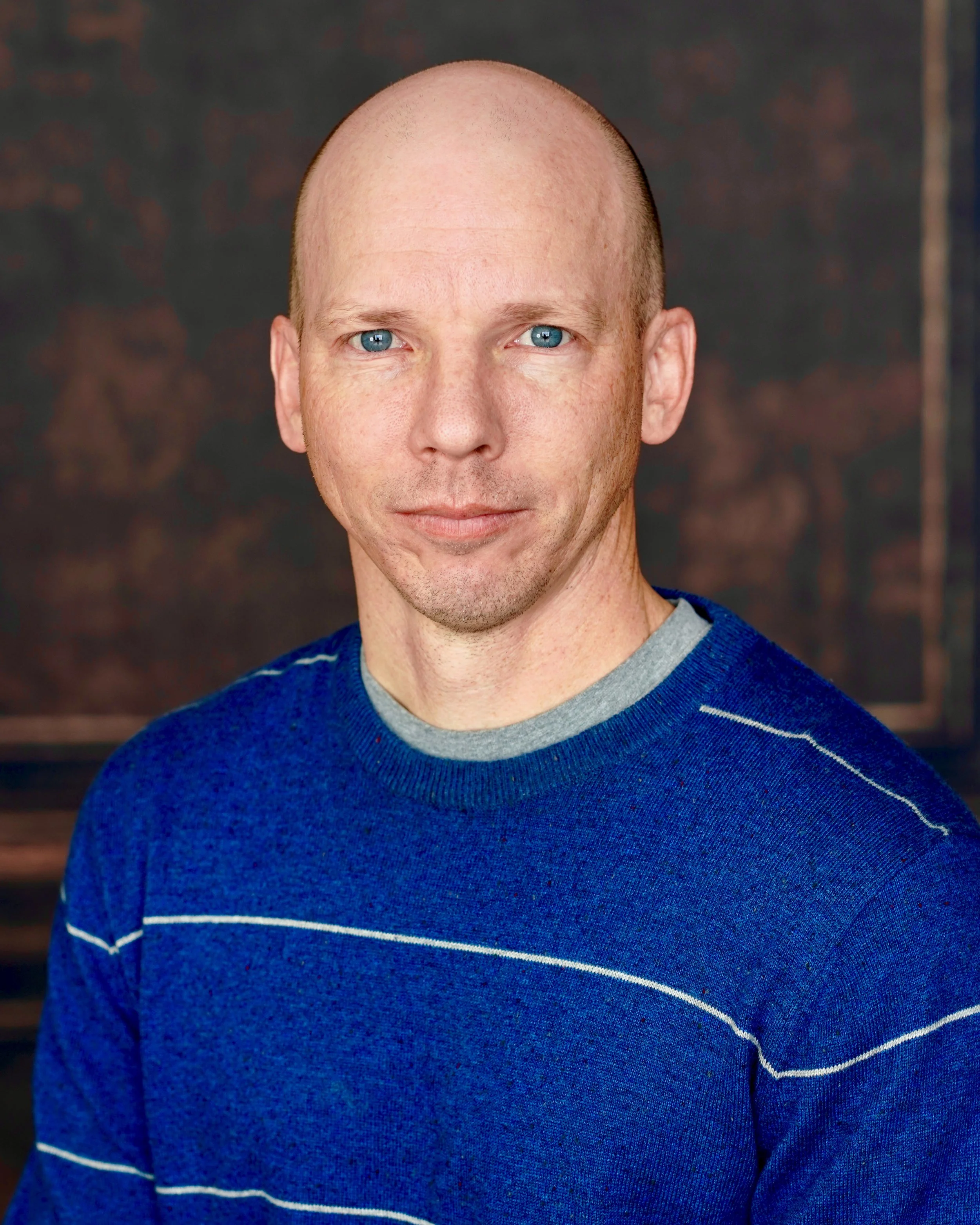 Headshot of a bald man with blue eyes, wearing a blue sweater with white horizontal stripes, standing against a dark, blurred background.