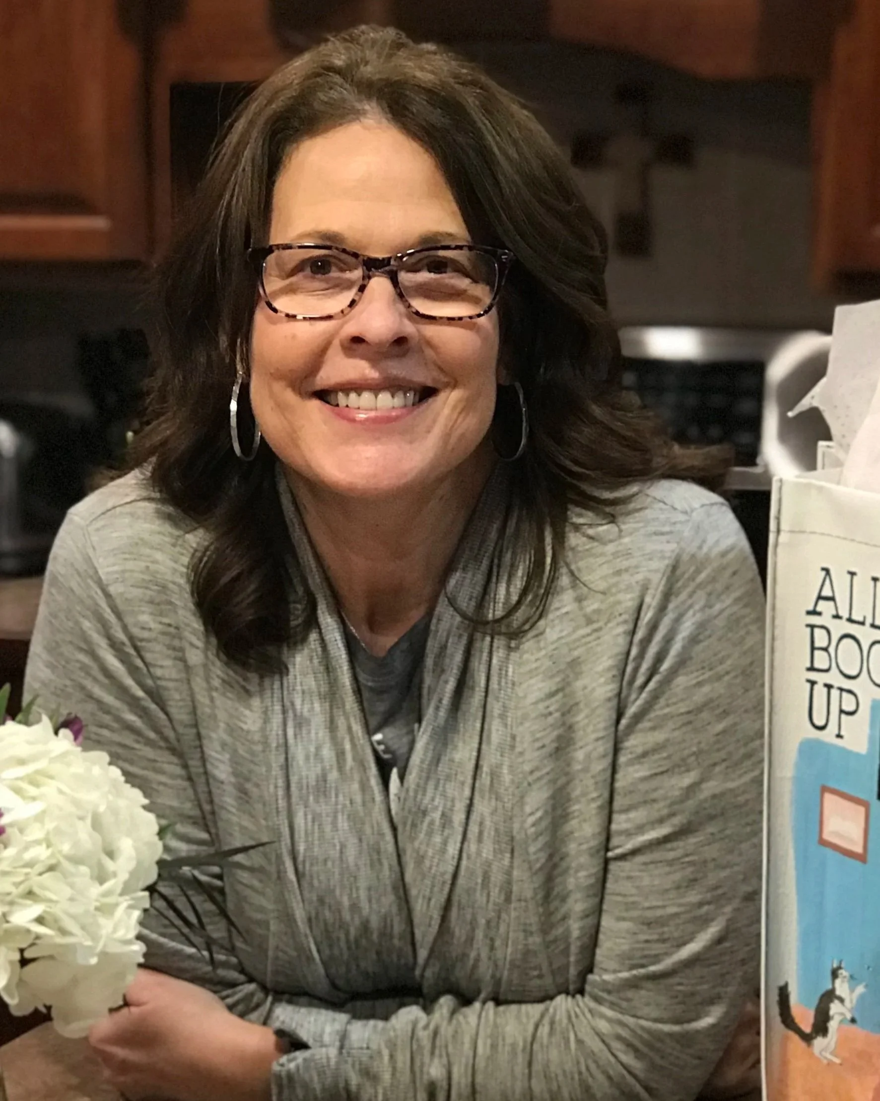 A woman with shoulder-length dark hair, glasses, and hoop earrings smiling at the camera. She is wearing a gray zip-up jacket and is seated at a table with a vase of white flowers. In the background, there is a kitchen with wooden cabinets and a microwave.
