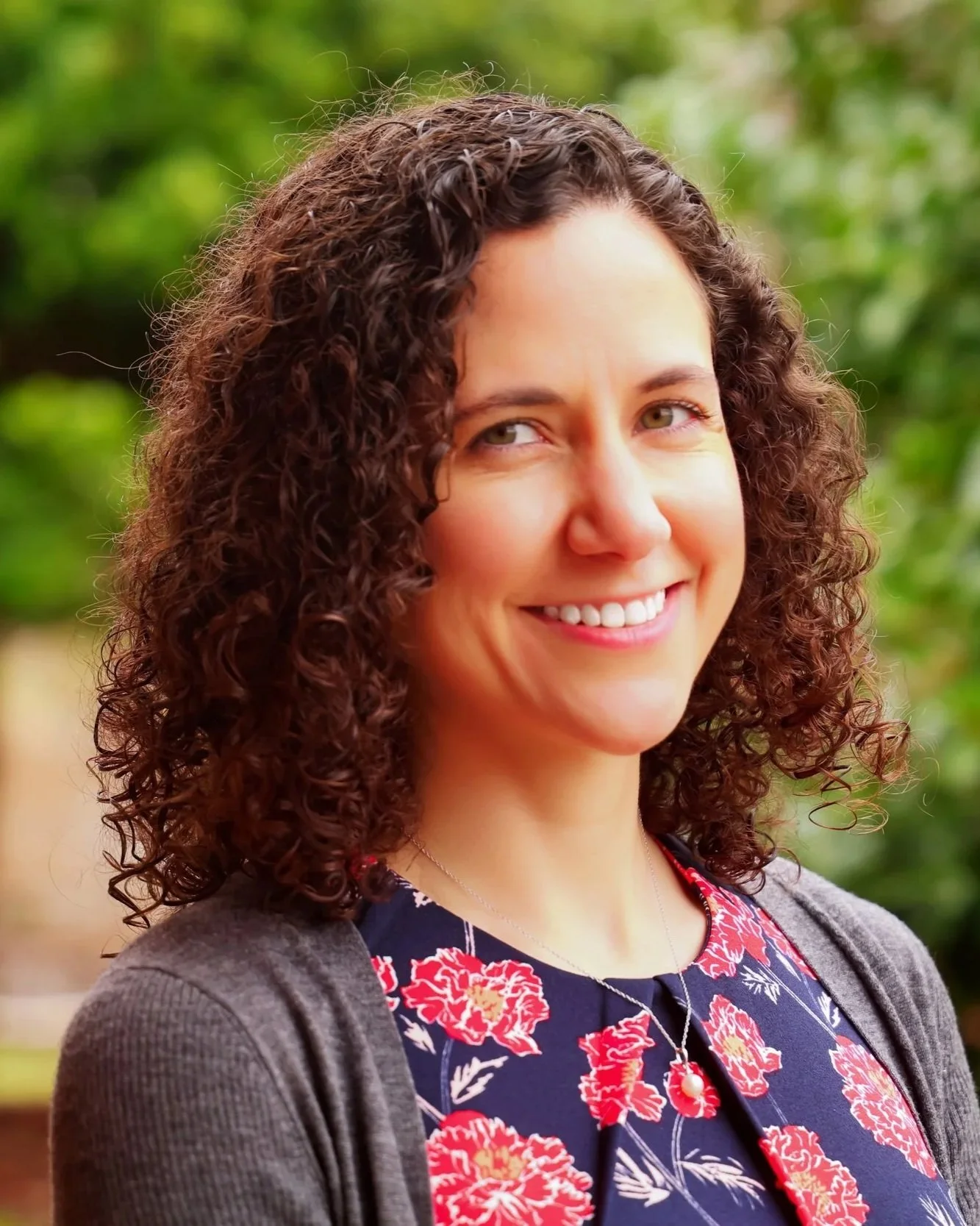 A woman with curly brown hair and light skin, smiling outdoors with blurred green foliage in the background.