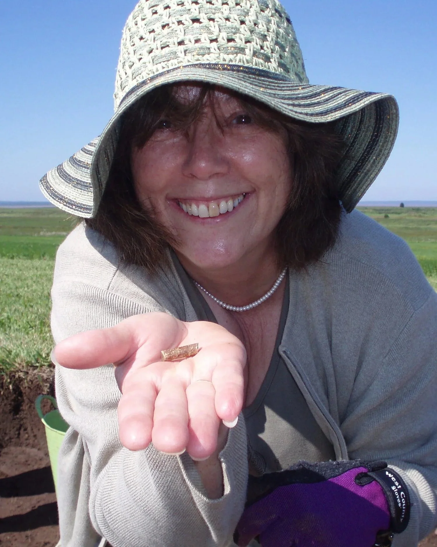 Smiling woman wearing a wide-brimmed hat and purple glove holds a small object towards the camera in an outdoor field with a clear blue sky