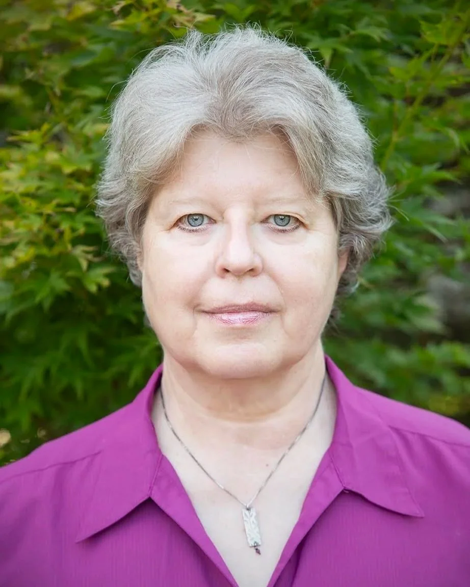 Portrait of an elderly woman with short gray hair and blue eyes, wearing a purple blouse and a silver necklace, standing outdoors with green foliage in the background.