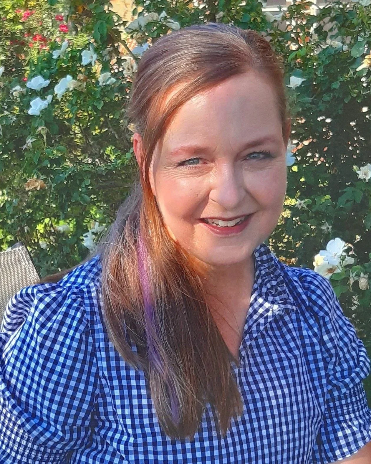 A smiling woman with long brown hair, wearing a blue and white checkered shirt, outdoors in front of a bush with white flowers.