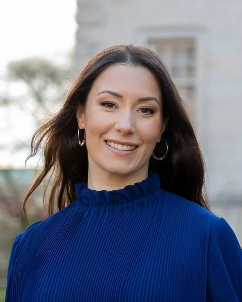 A woman with long brown hair wearing a blue ruffled top and hoop earrings, smiling outdoors with a blurred building and trees in the background.