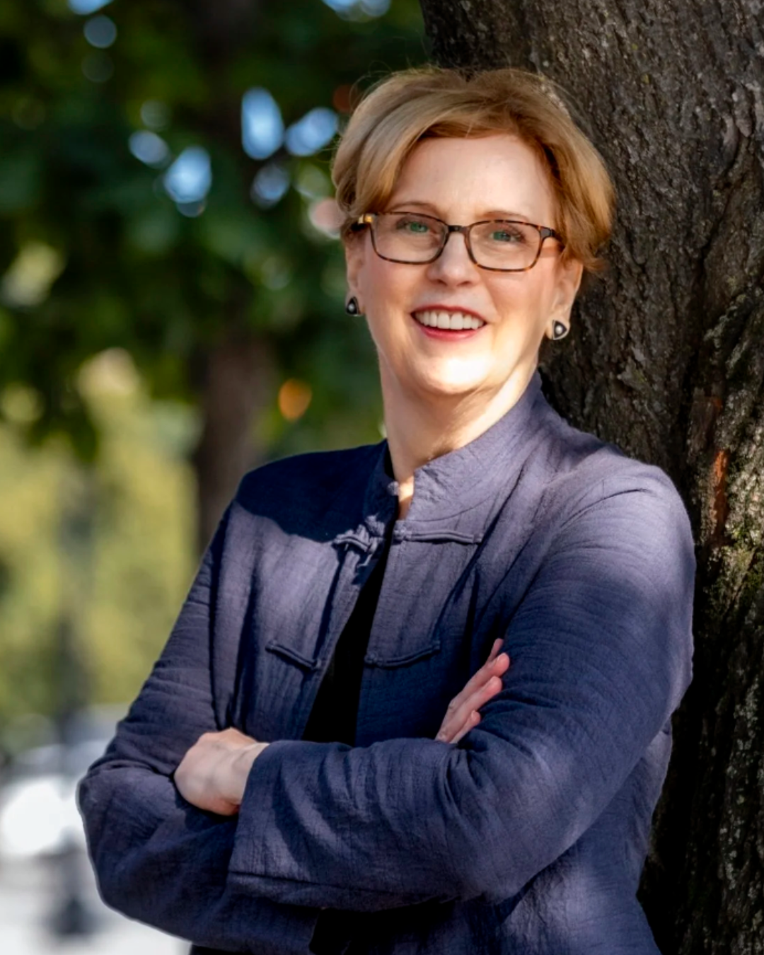 A woman with short blonde hair and glasses smiling, leaning against a tree outdoors.