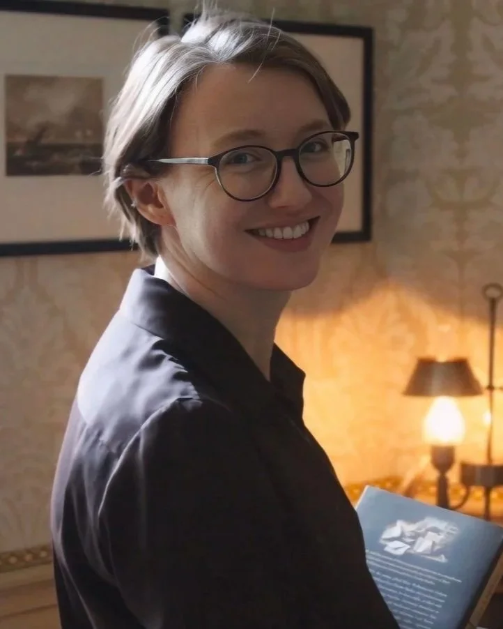 A smiling woman with short gray hair and glasses, wearing a dark shirt, holding a book in a warmly lit room.
