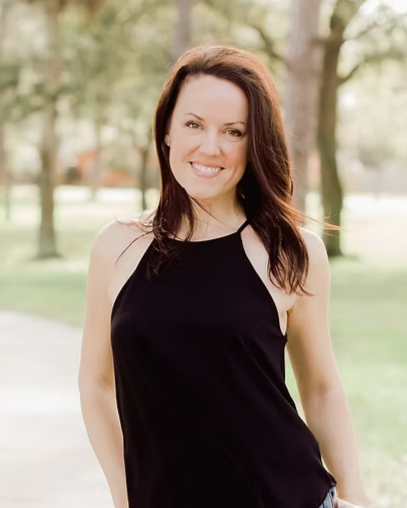 A woman with dark brown hair smiling outdoors in a park with trees and grass.