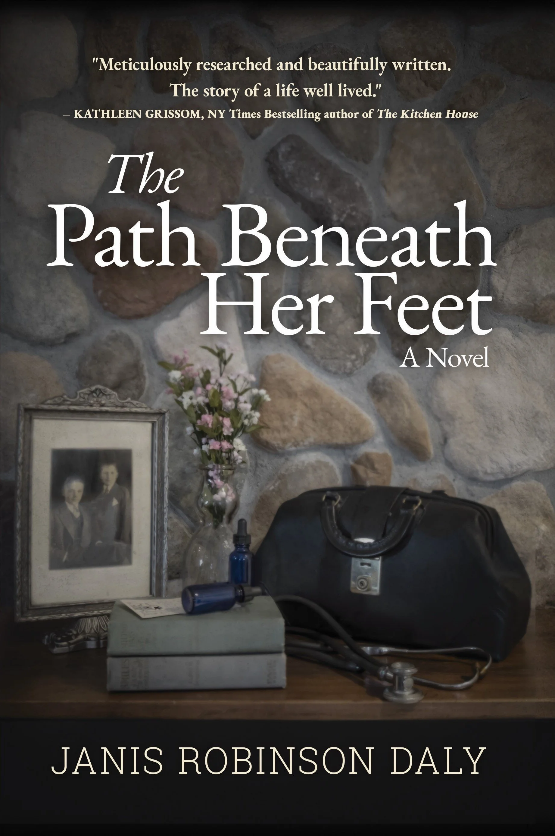 Book cover for "The Path Beneath Her Feet" by Janis Robinson Daly, featuring a stone wall background, a framed black-and-white photo, a glass vase with pink flowers, a stethoscope, a dark medical bag, and a bottle of eye dropper medicine on a wooden surface.