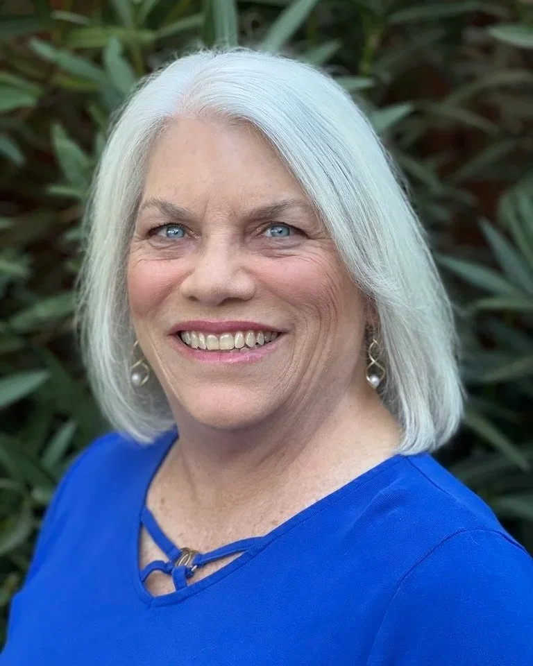 A smiling woman with shoulder-length white hair and blue eyes, wearing a royal blue top and pearl earrings, outdoors with green foliage in the background.
