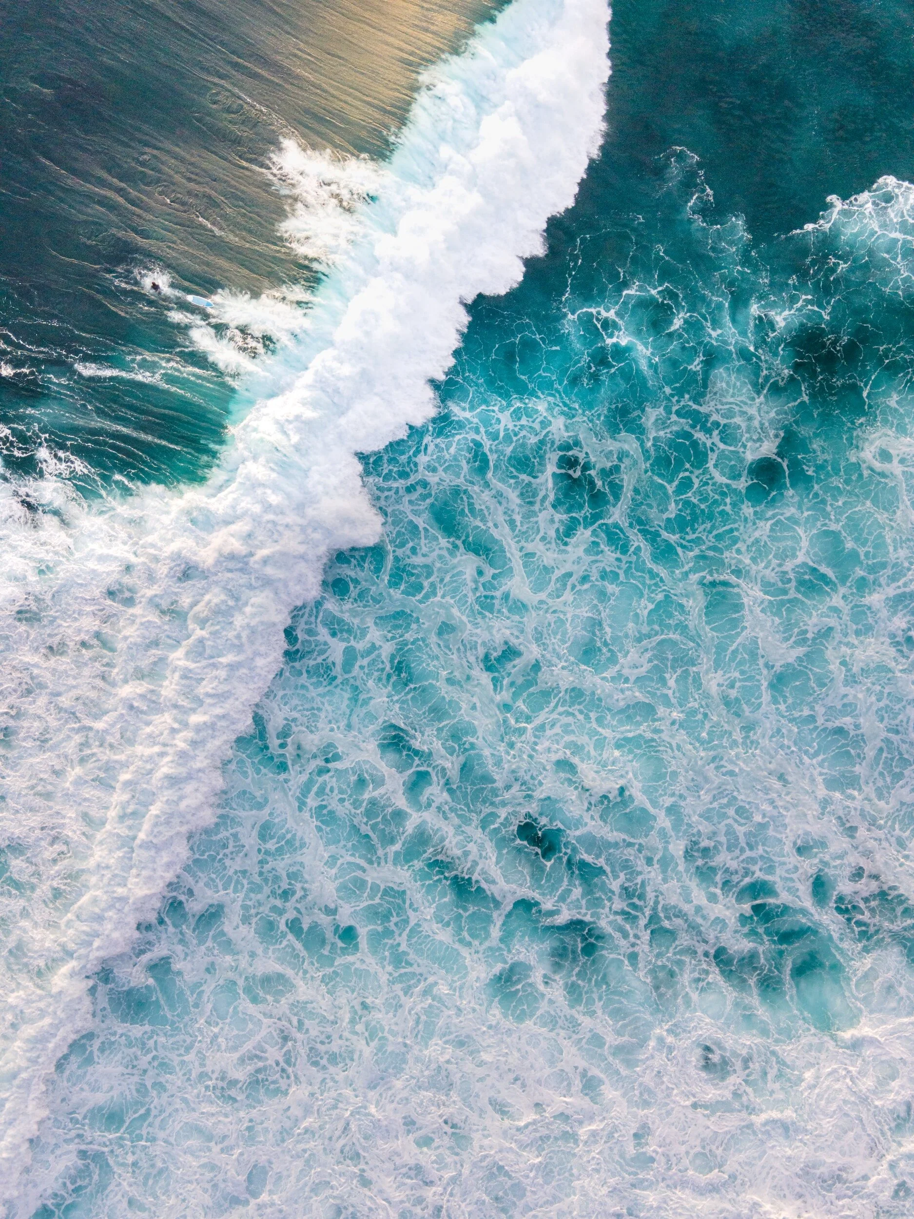 Aerial view of ocean waves crashing onto the shore, creating white foam and swirling patterns in turquoise water.