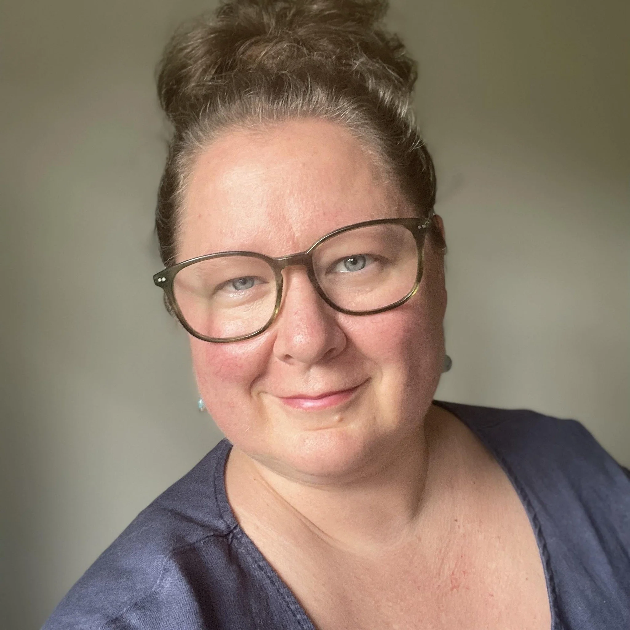A close-up of a woman wearing a dark blue top, brown hair in a bun, and black glasses.