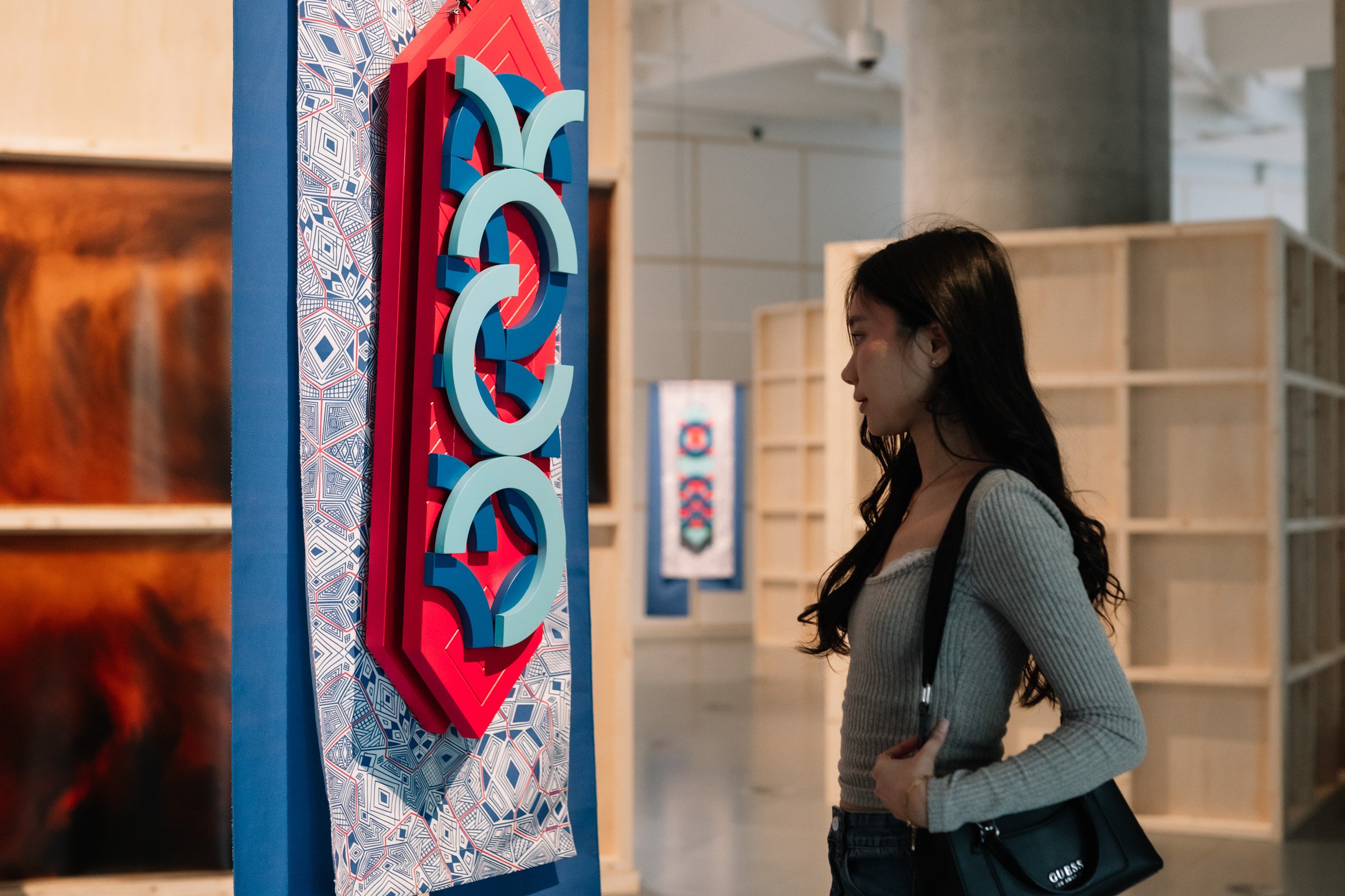 A woman with long dark hair and a grey long sleeve looking at a colourful hanging sculptural installation piece.