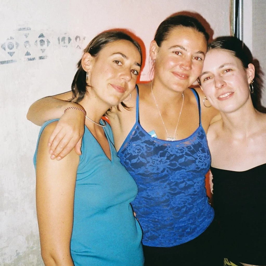 A person with shoulder length brown hair and a teel tank top, a person with brown hair tied up and a blue lace tank top and a person with brown hair tied up in a black tank top standing together in a side hug