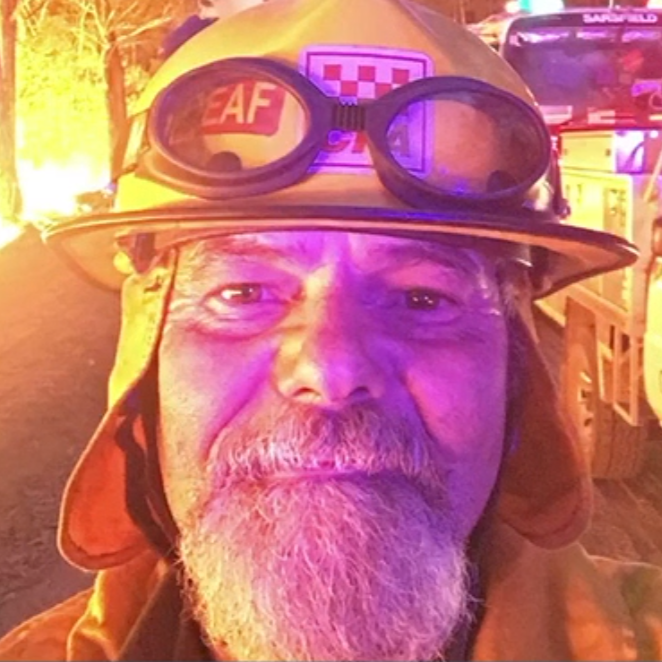 A close-up photo of a man with a beard in a fire fighter gear, standing in front of a bright flame blaze.