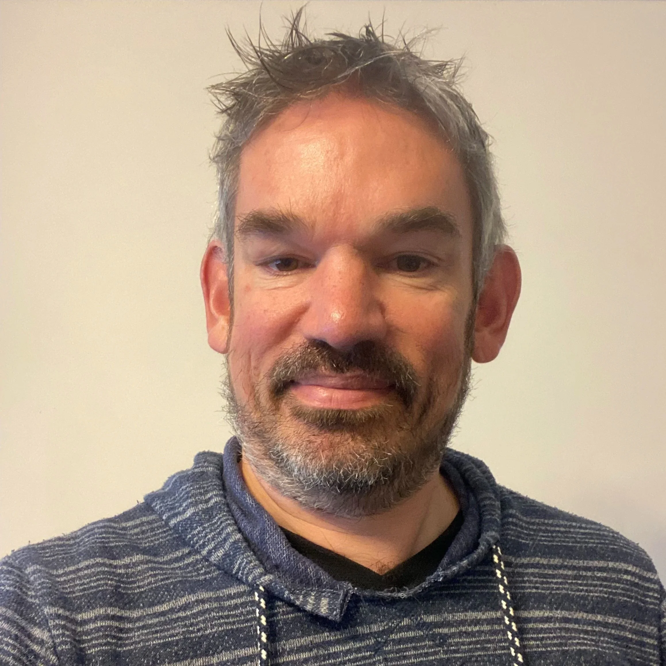 A close up of a man with grey hair and facial hair, wearing a striped grey jumper.