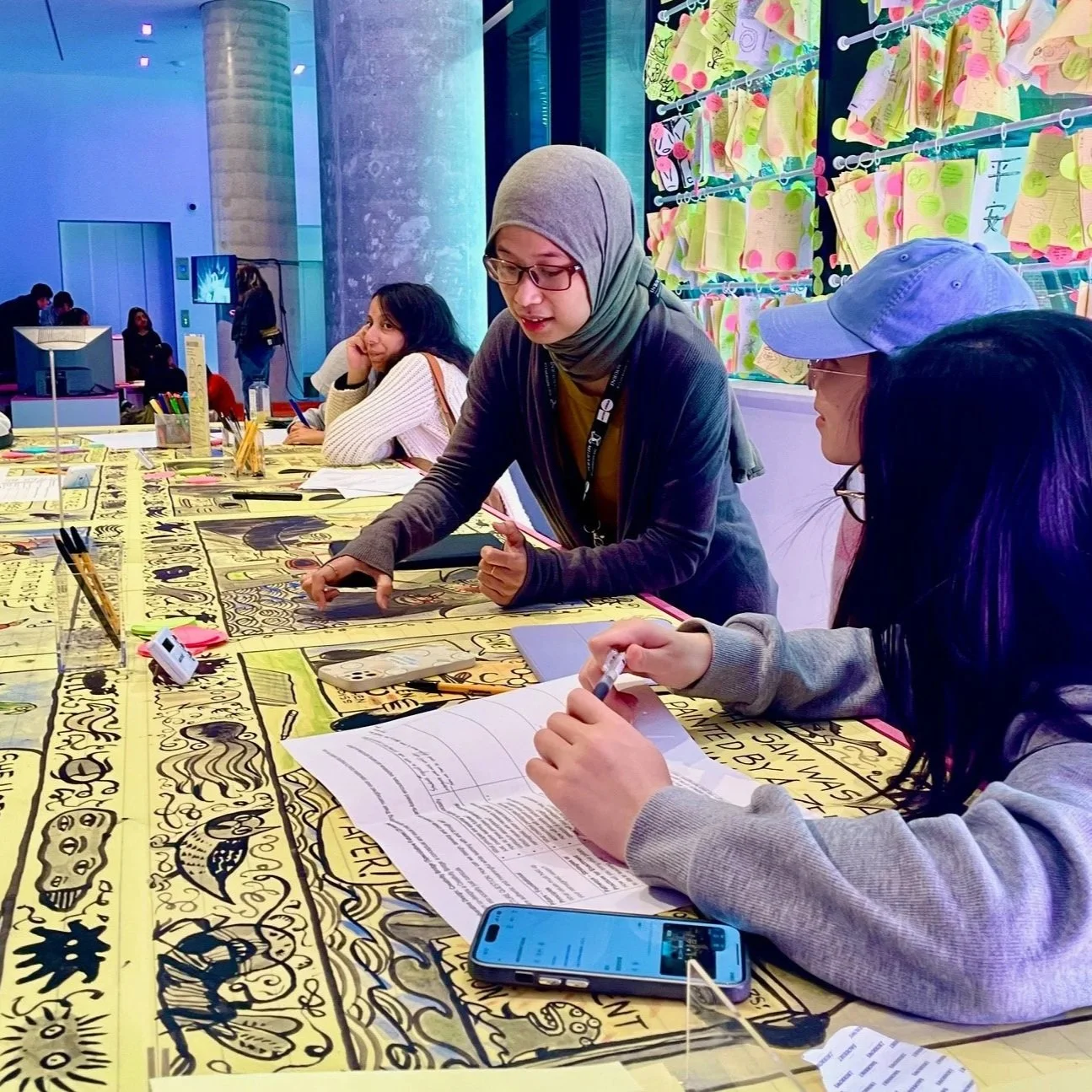 A student works with two other students on a colourful table with hand drawn artwork