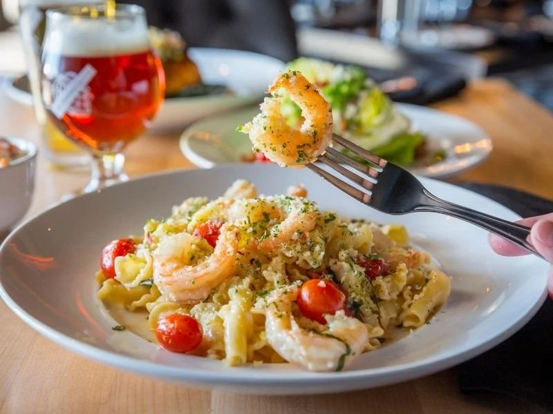 A fork lifting a piece of shrimp and pasta from a bowl of seafood pasta, with cherry tomatoes and grated cheese, on a wooden table with a glass of beer in the background.