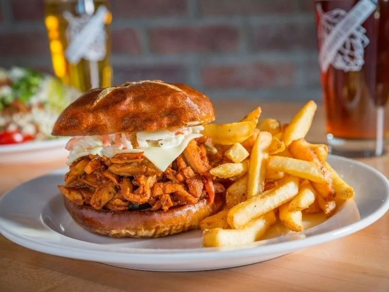 Pulled pork sandwich with coleslaw and a side of French fries on a white plate, with a glass of beer in the background.