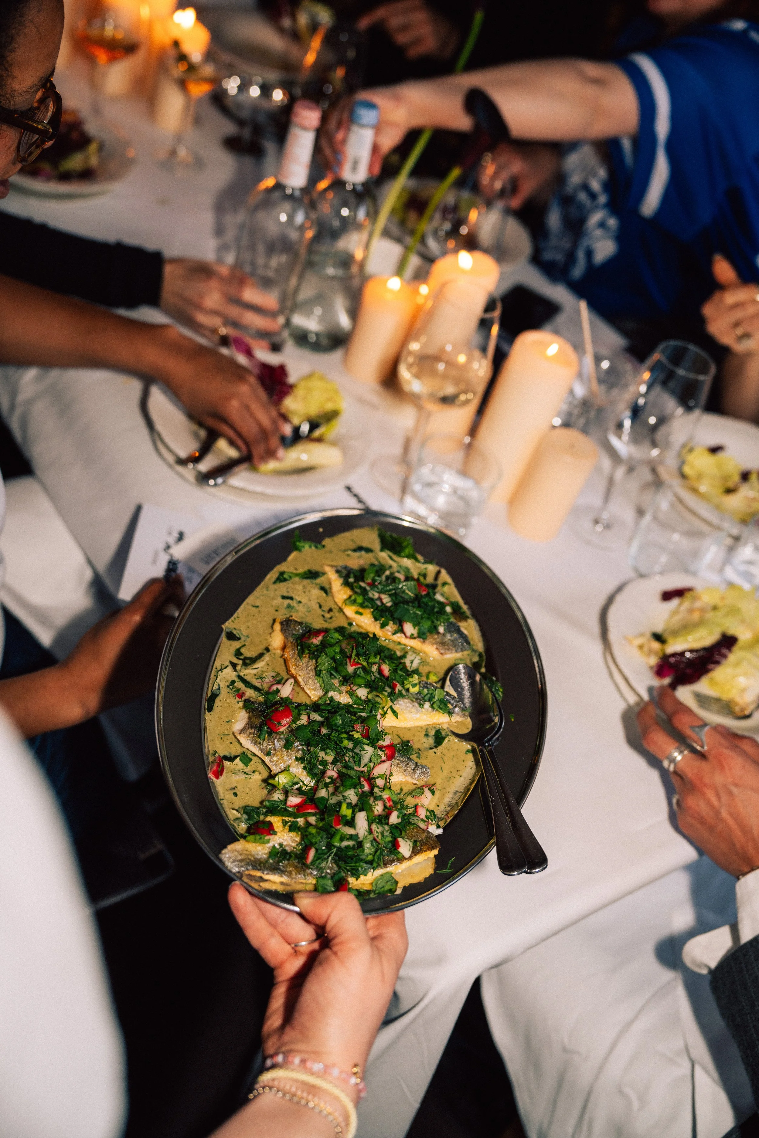 Person holding a plate of fish garnished with herbs and radish slices at a dinner table with candles and wine bottles.