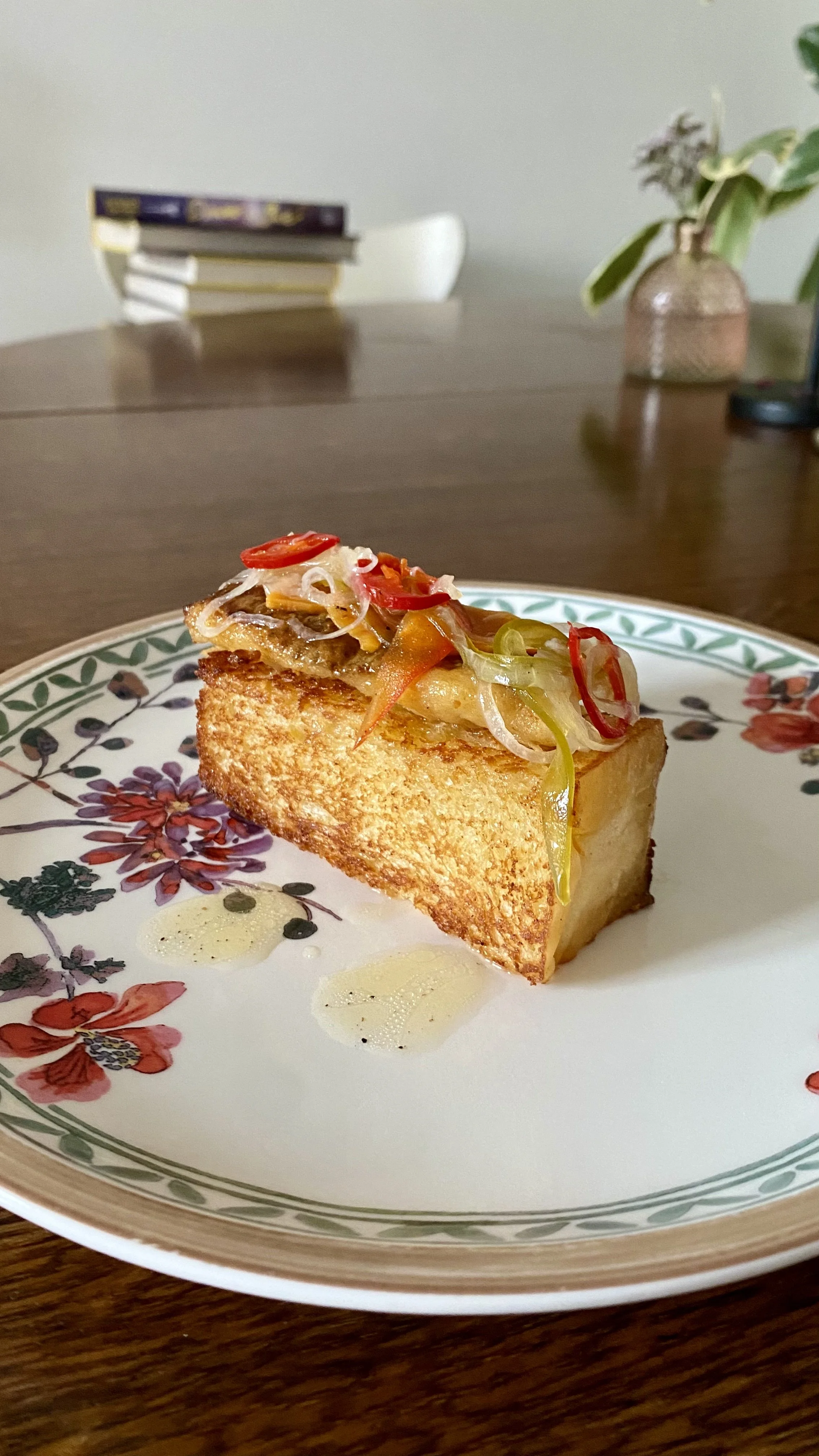 A slice of pie with a golden crust on a floral plate, topped with shredded vegetables and a drizzle of sauce, set on a wooden table.
