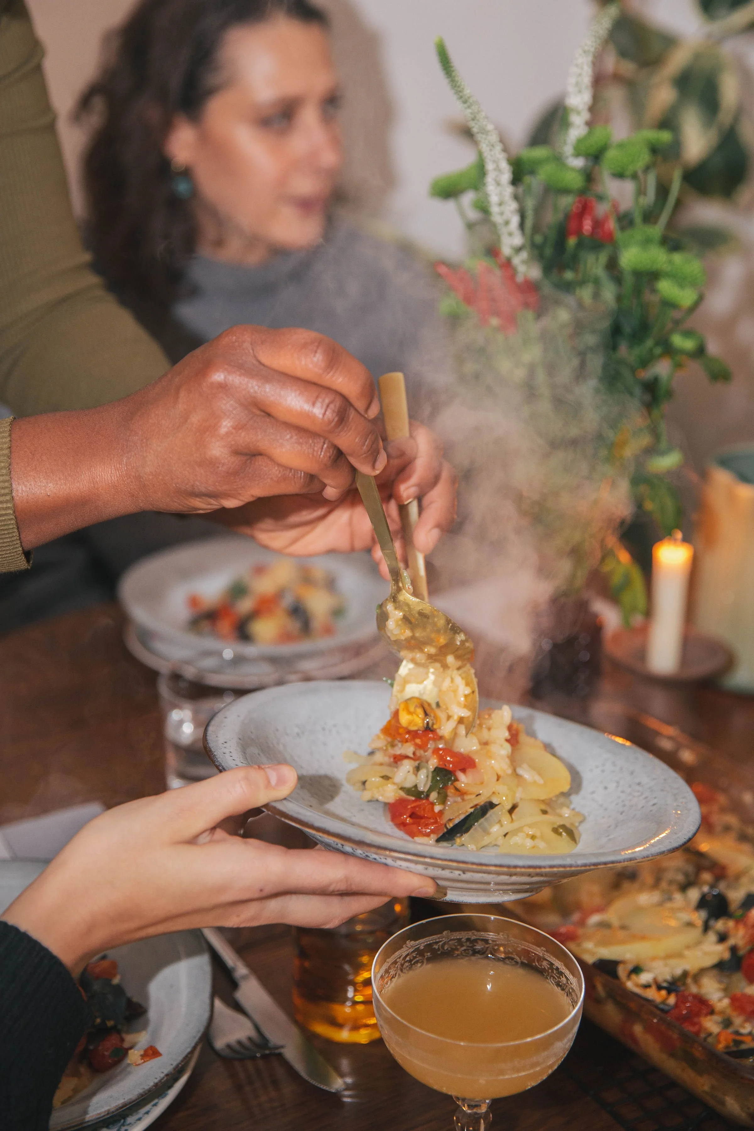 A person with dark skin is holding a plate of pasta with vegetables, while someone else uses chopsticks to serve the pasta onto the plate. In the background, there is a woman with curly hair and earrings, and a table with dishes, a lit candle, and a 