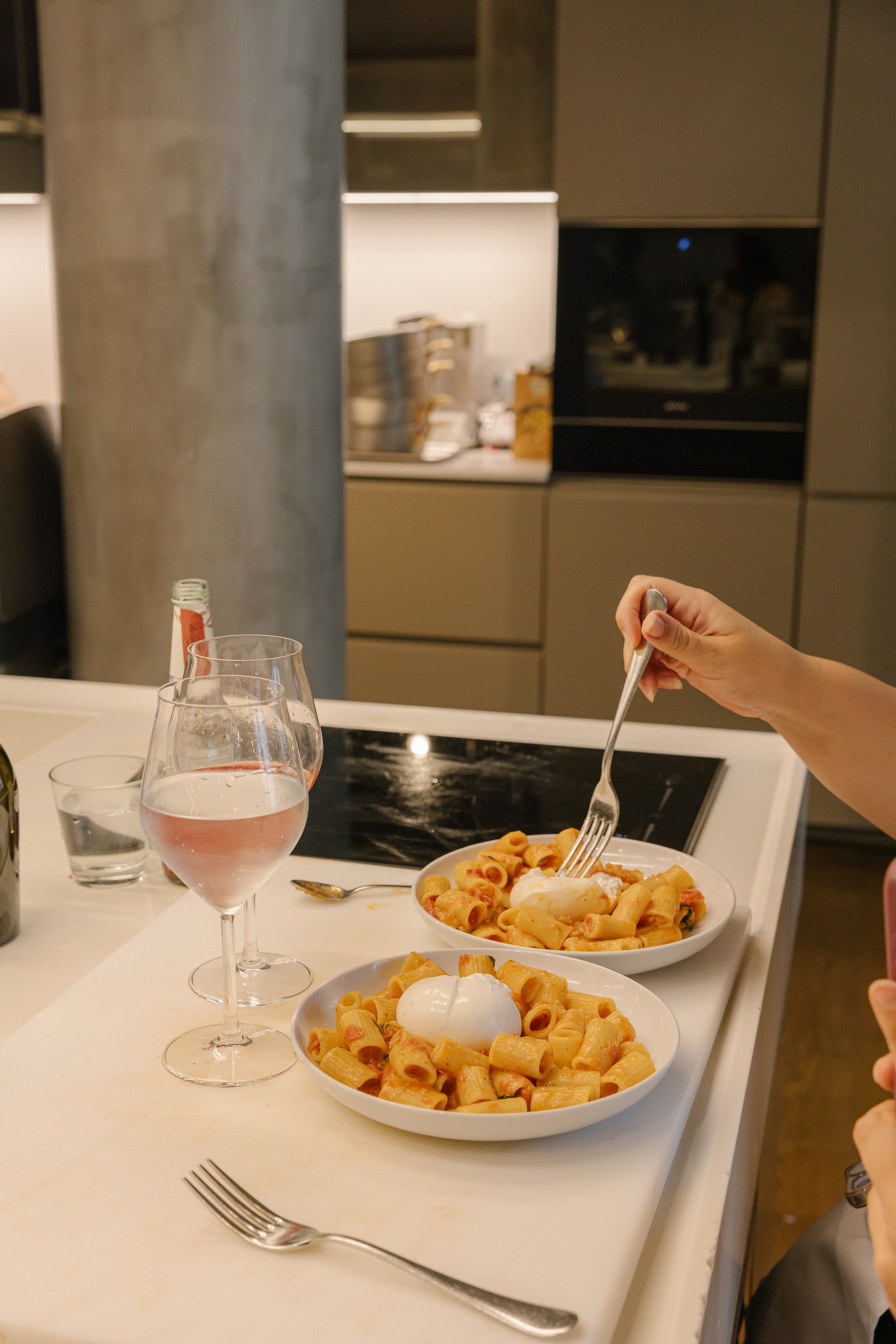 A dinner scene with two bowls of pasta topped with poached eggs, a glass of water, and a glass of rosé wine on a white table. A hand holding a fork is reaching into the pasta. The background shows kitchen cabinets and a person reflected in a mirror.