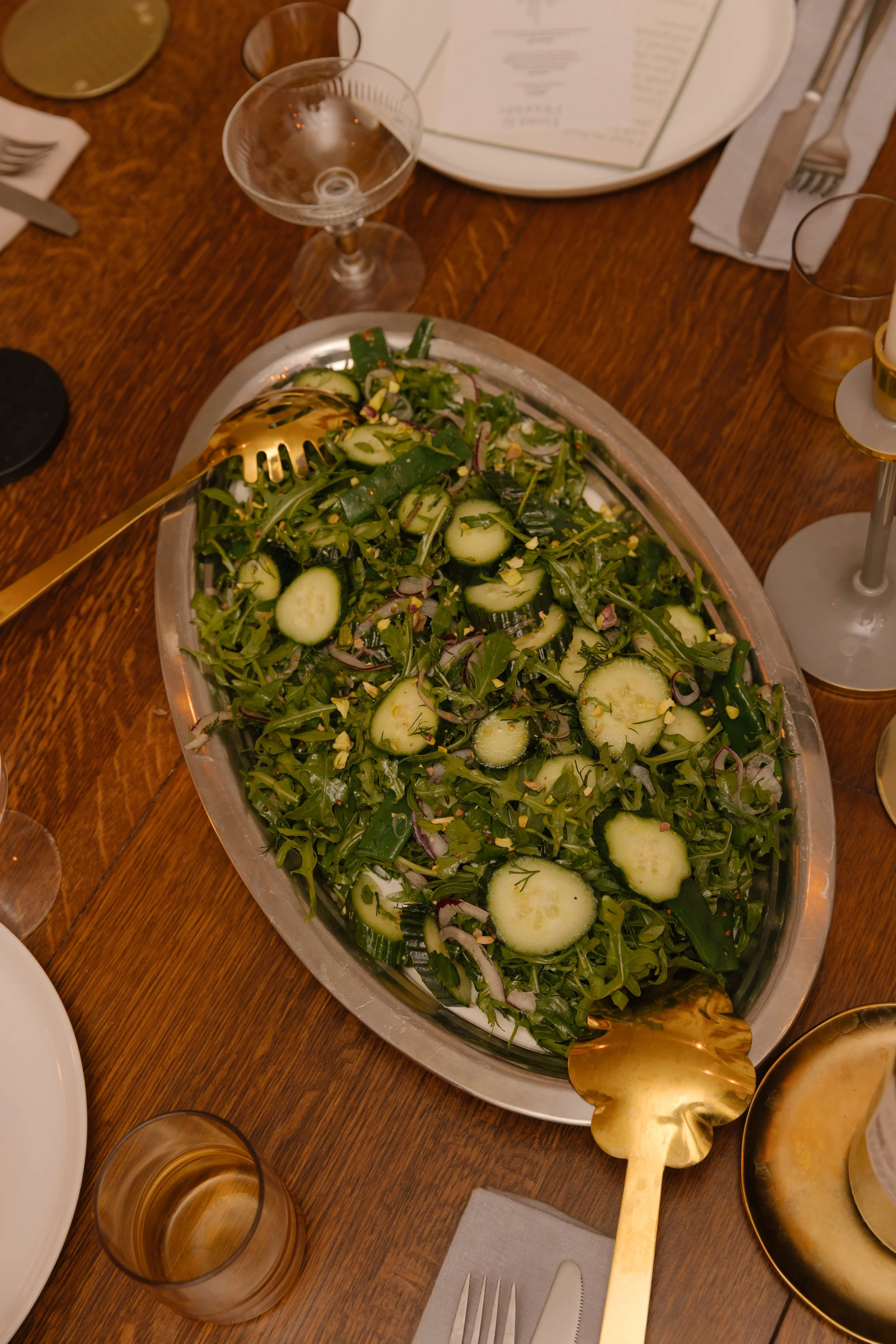 A large silver tray of cucumber salad with chopped herbs and red onions on a wooden dining table. Surrounding the tray are empty glasses, plates, utensils, and a menu on the table.