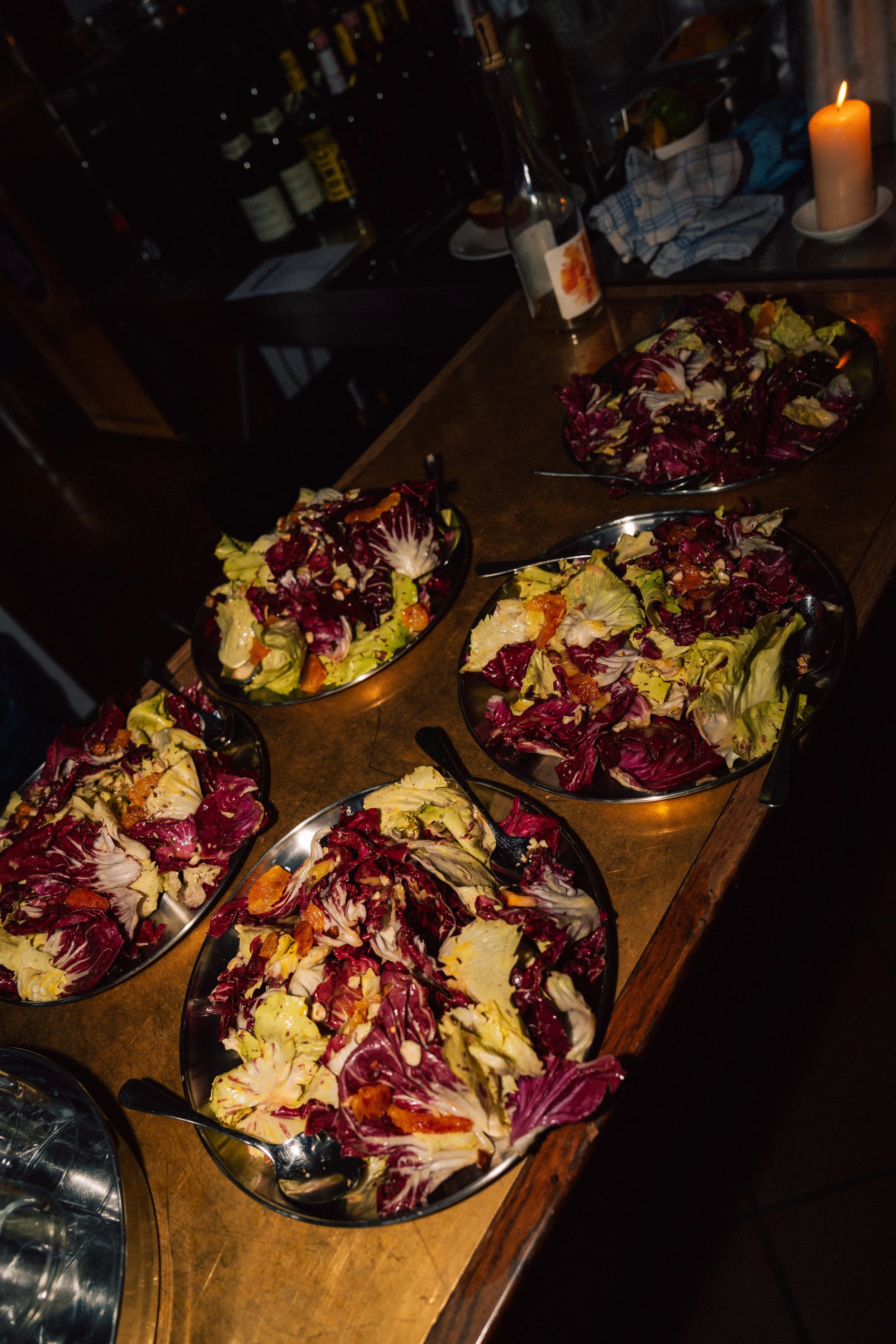 Several bowls of colorful salad with lettuce and vegetables on a wooden table, with a lit candle and bottles in the background.