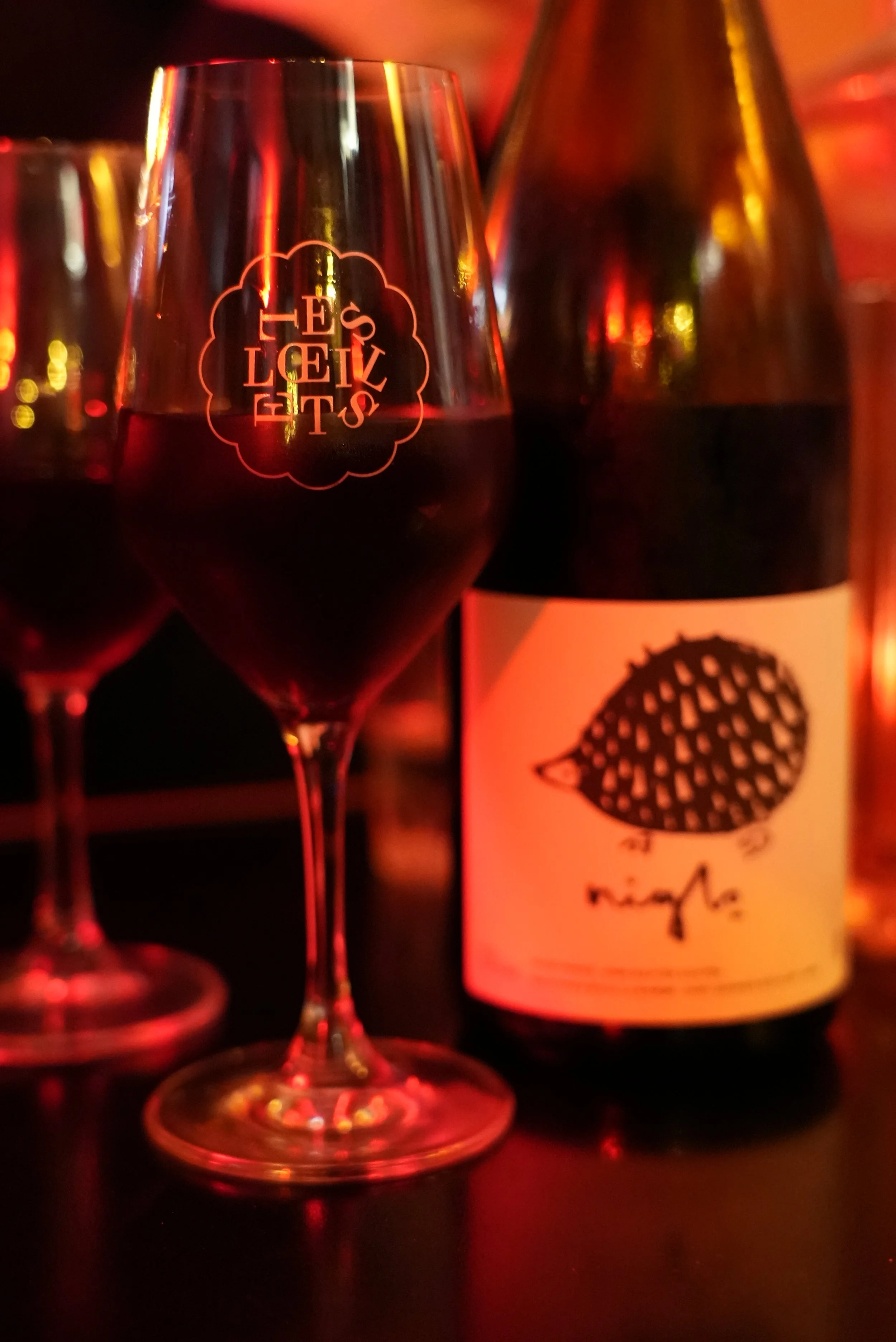 A glass of red wine with a logo that says 'Les Oeillets" and a wine bottle behind it with a black and white label featuring an illustration of a hedgehog.