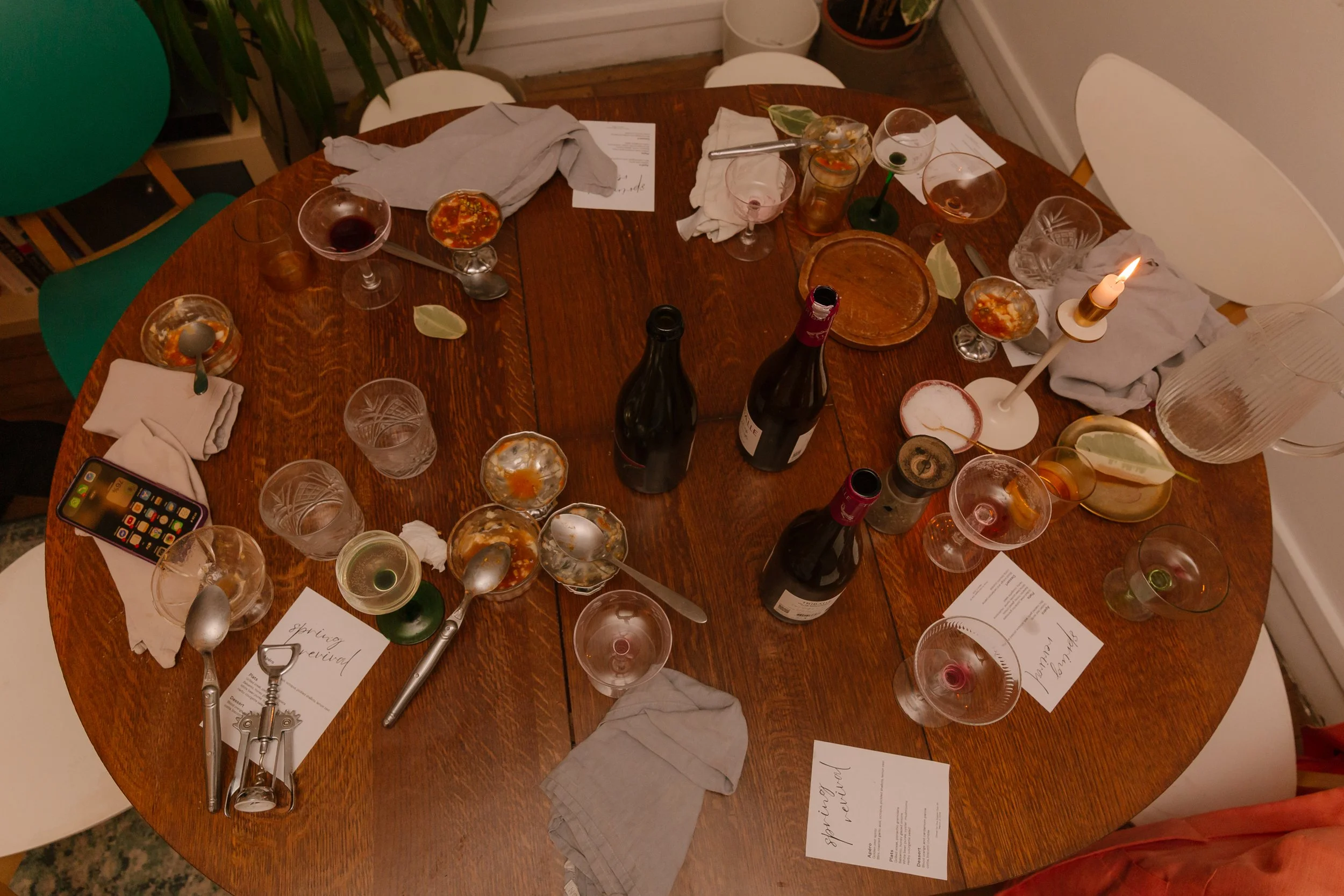 A wooden dining table with several empty and used glasses, bottles of wine, scattered paper notes, a candle, a smartphone, and crumpled napkins in a casual setting.