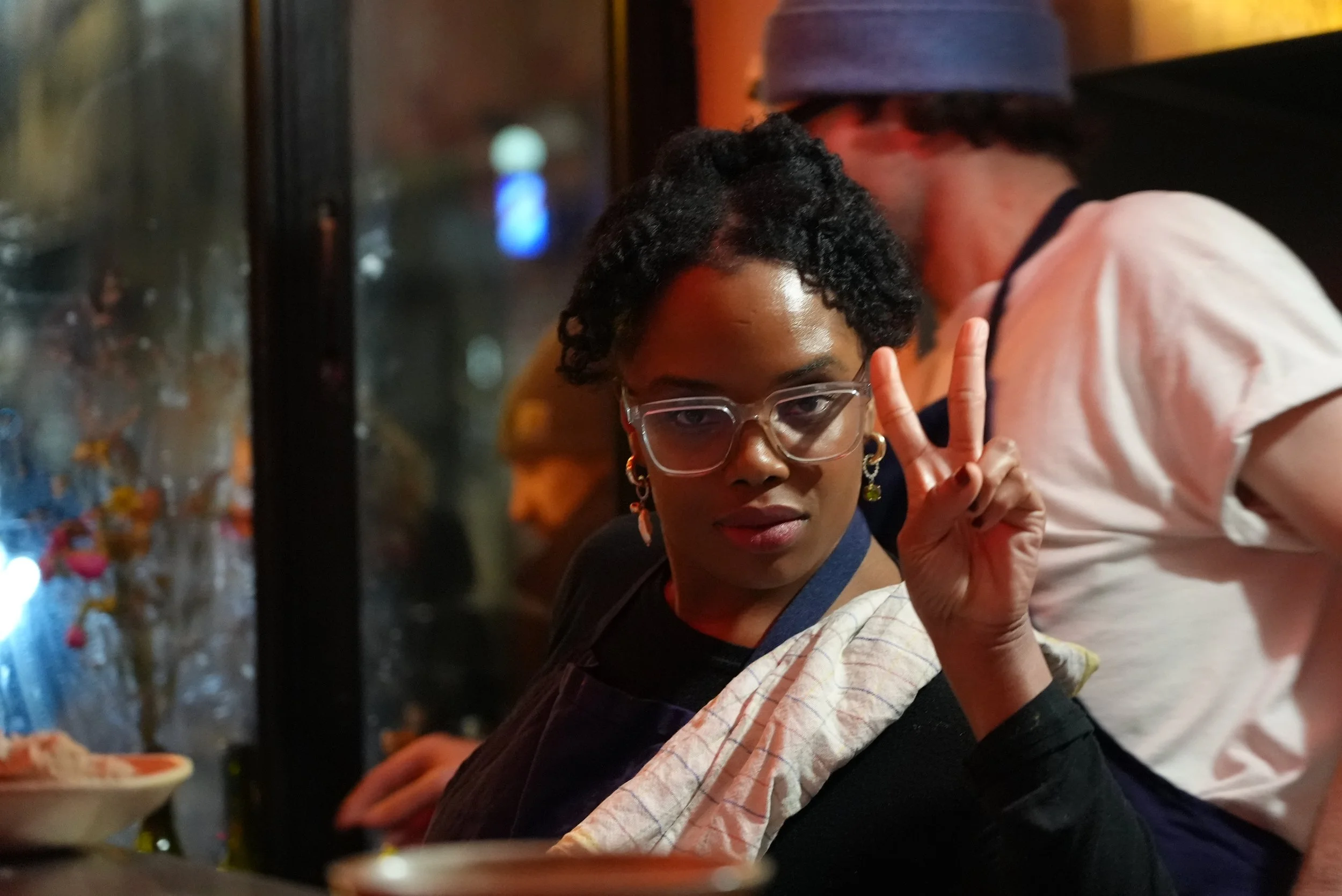 A young person with short, curly hair and glasses making a peace sign gesture in a dimly lit setting, possibly a restaurant or bar, with blurry colorful lights and a woman sitting nearby in the background.