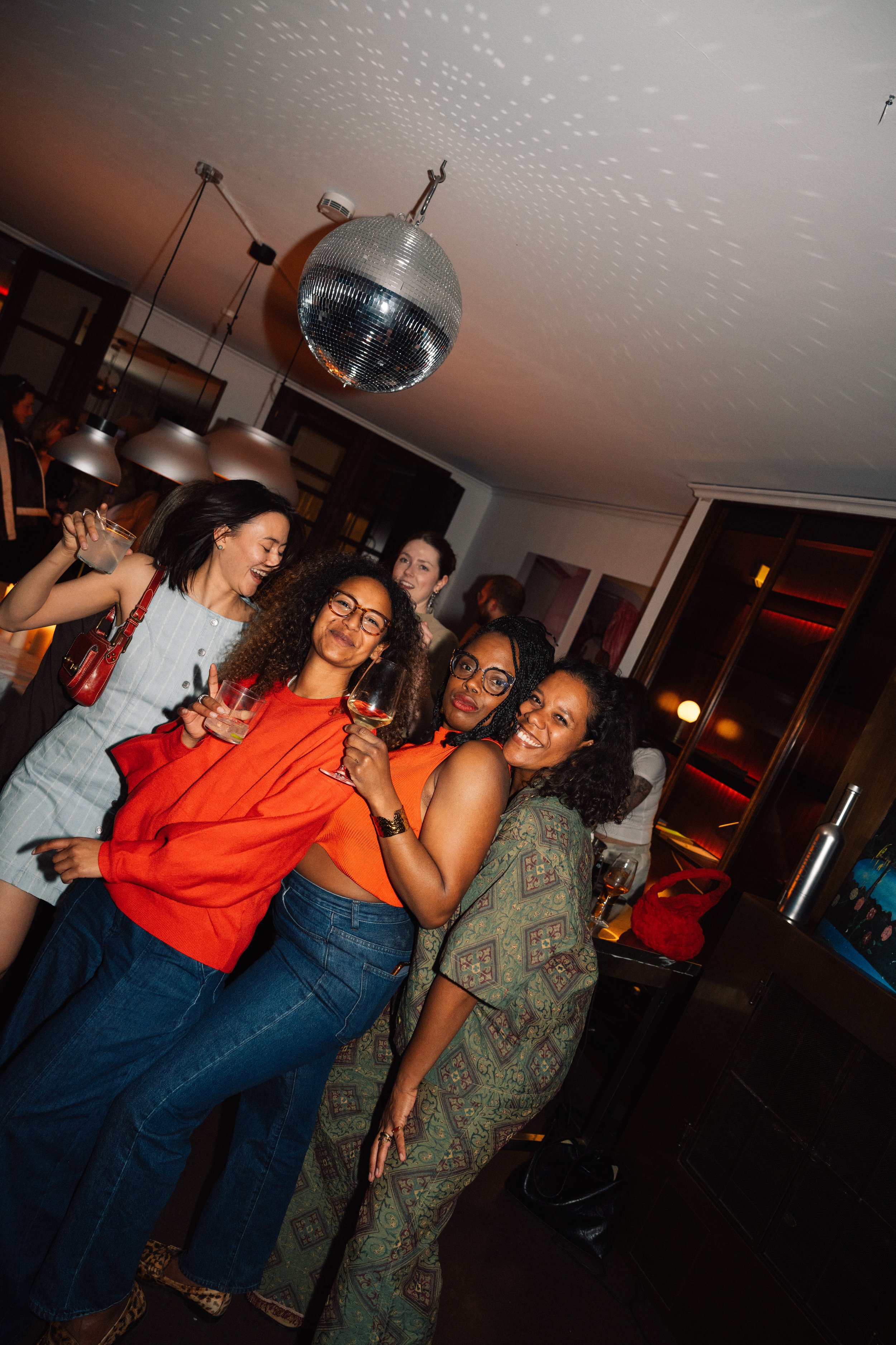 Five women at a bar holding drinks and smiling, with a disco ball casting light patterns on the ceiling in a warmly lit indoor setting.