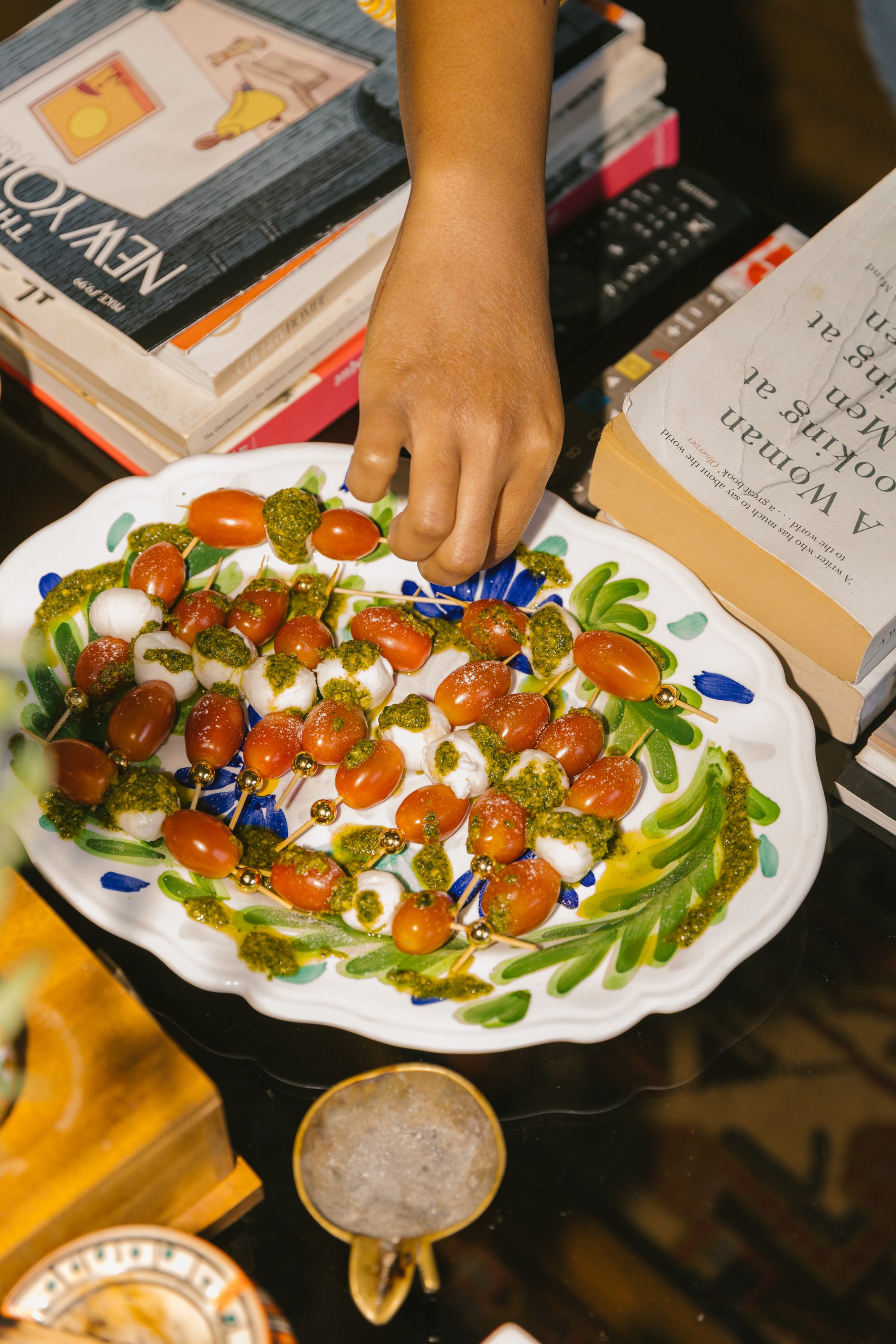 A person reaching for cherry tomatoes and mozzarella balls on a decorative plate, surrounded by various books, a remote control, and bowls of salt and spices on a dark surface.