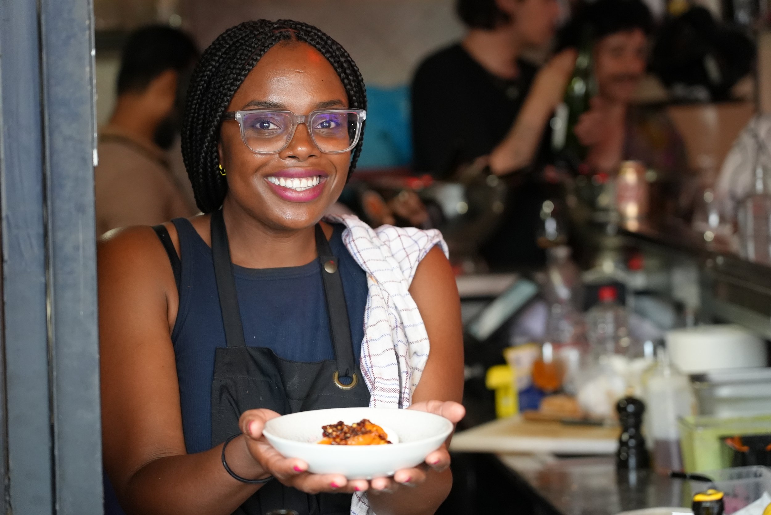 Smiling woman with braids and glasses holding a bowl of food in a kitchen or restaurant setting.