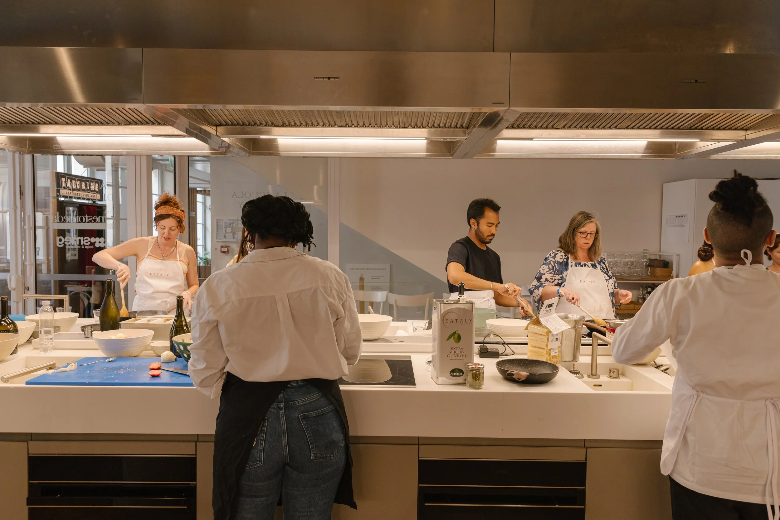 People cooking in a professional kitchen with stainless steel appliances and a white counter, some wearing aprons and others dressed casually.