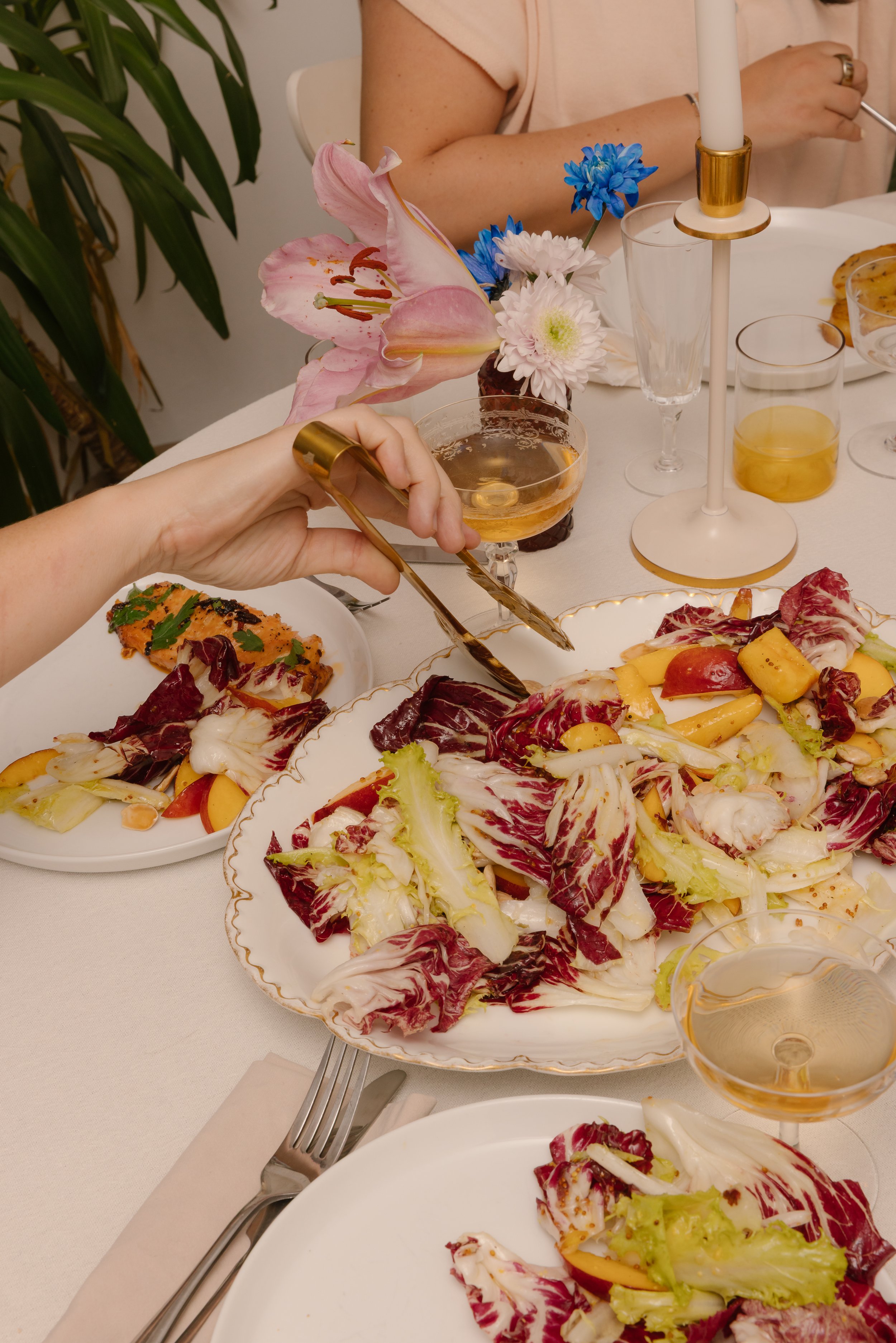 A table set for a meal with plates of salad, a floral centerpiece, empty champagne glasses, and a lit candlestick. A person is reaching for food with tongs.