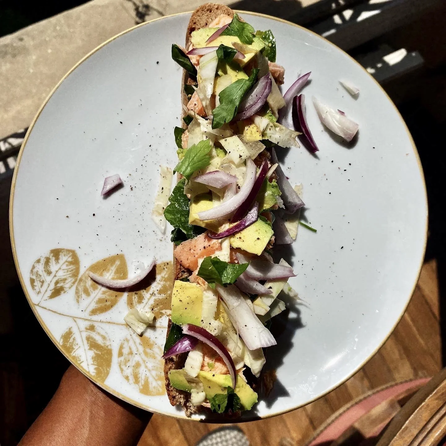 A slice of bread topped with chopped avocado, lettuce, onion, and herbs, served on a decorative white plate with a gold floral pattern.