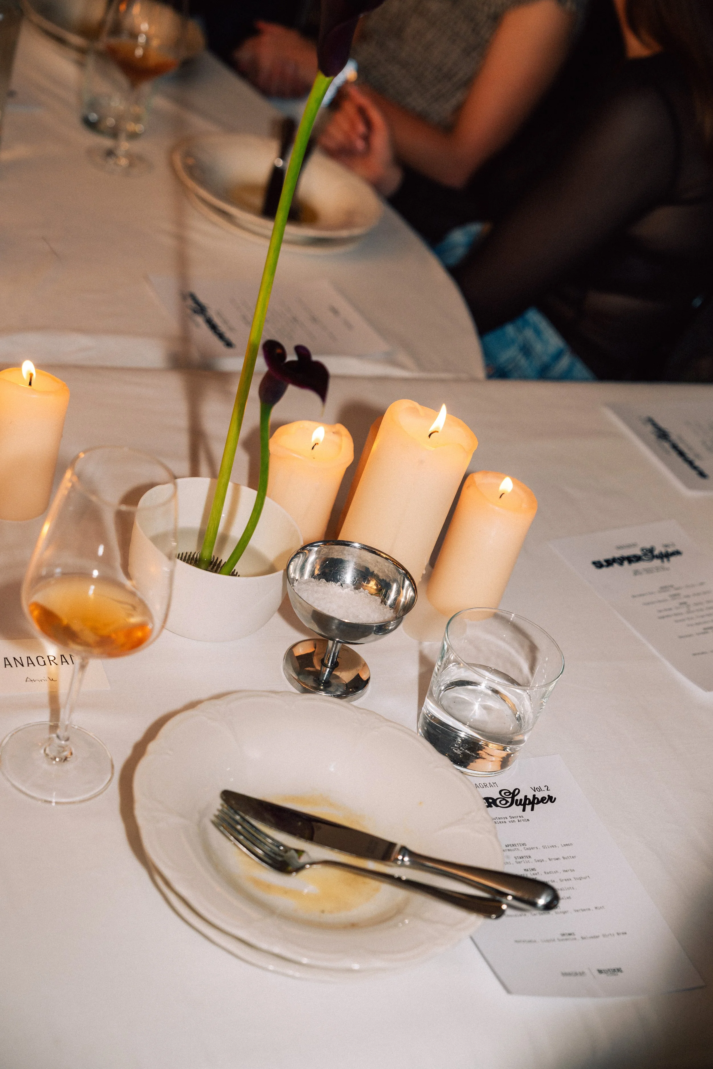 A table setting with a white plate, silverware, a half-filled glass of water, a wine glass with some amber-colored drink, and several lit candles. There are also menus and a vase with dark flowers, along with some people in the background.