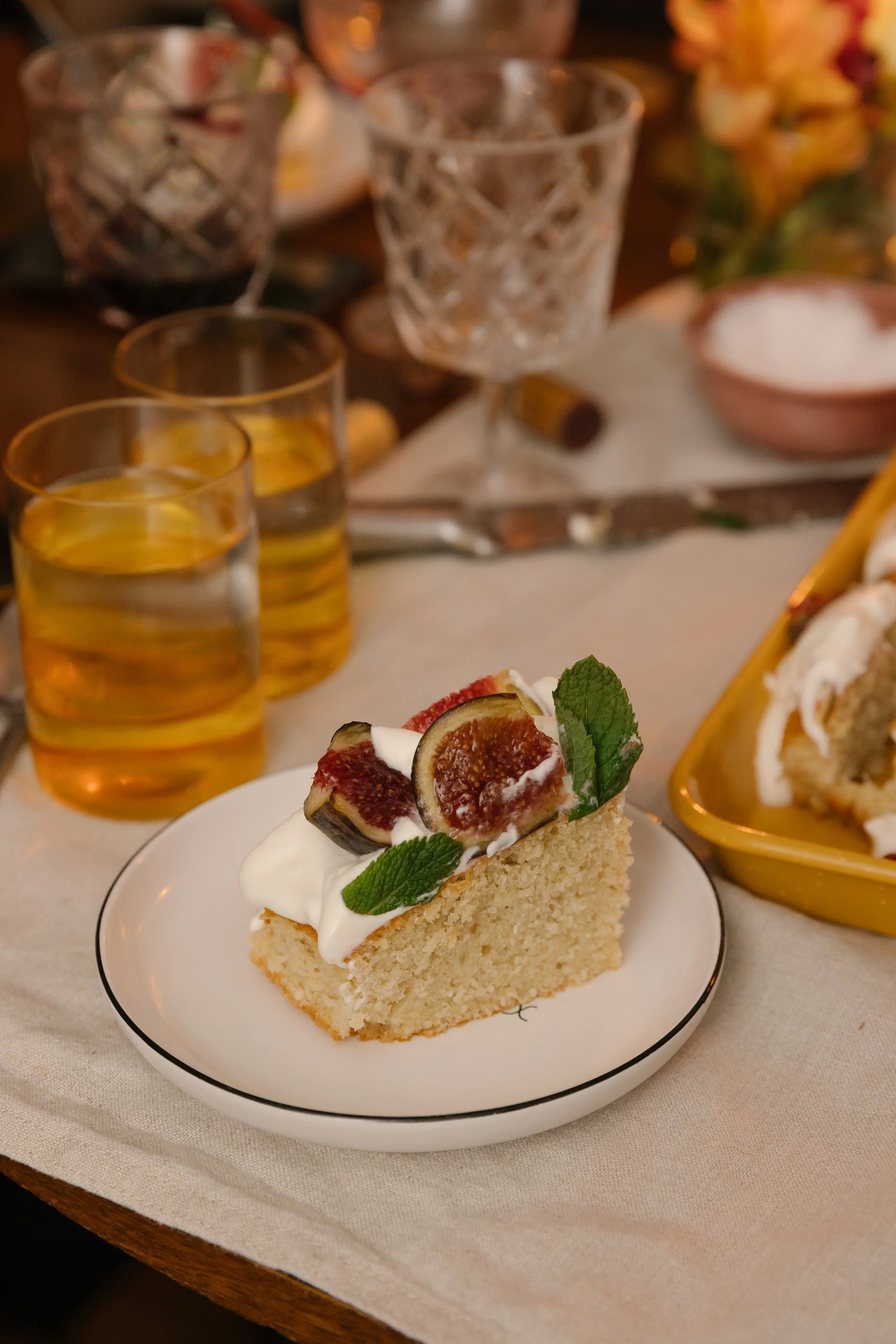 A slice of vanilla cake topped with whipped cream, fresh figs, and mint leaves on a white plate.